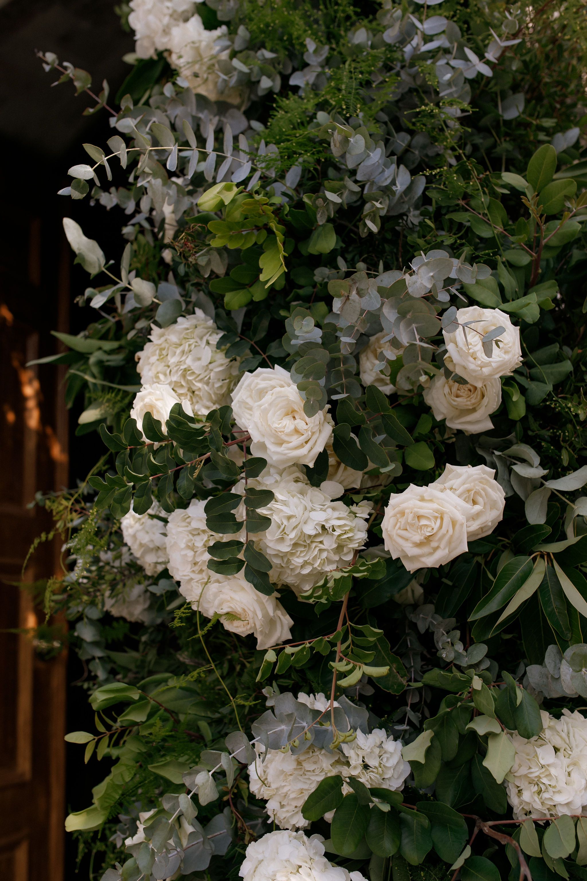 Cream-white garden roses and white hydrangea heads are woven through a dense wall of green foliage, with silvery dollar eucalyptus and glossy leaves scattered among them, forming a lush wedding floral installation in Greece. The blooms cluster in soft, rounded groups, and their pale petals stand out against the deep greens. Warm, low light falls on the flowers from the left, beside a wooden door in the shadows, giving the arrangement a romantic, understated feel.