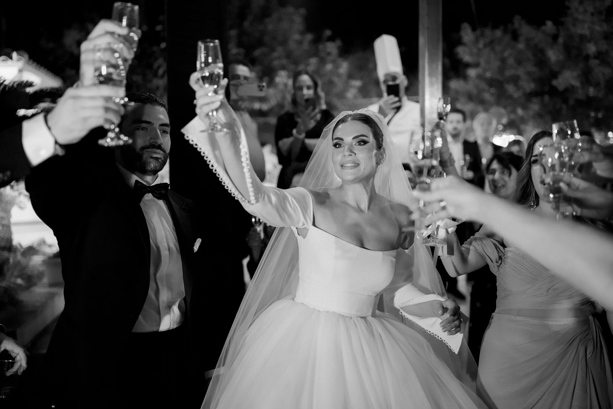 A black and white night photo of a bride in a fitted strapless gown with a full tulle skirt and a long veil, raising a champagne flute high in her right hand as she smiles toward the crowd. Beside her, a groom in a black tuxedo and bow tie also lifts a glass, while guests around them raise their own glasses and hold up phones. This moment of a toast at a wedding in Greece was captured at a reception, and the blurred trees and terrace behind the couple suggest an evening outdoors.