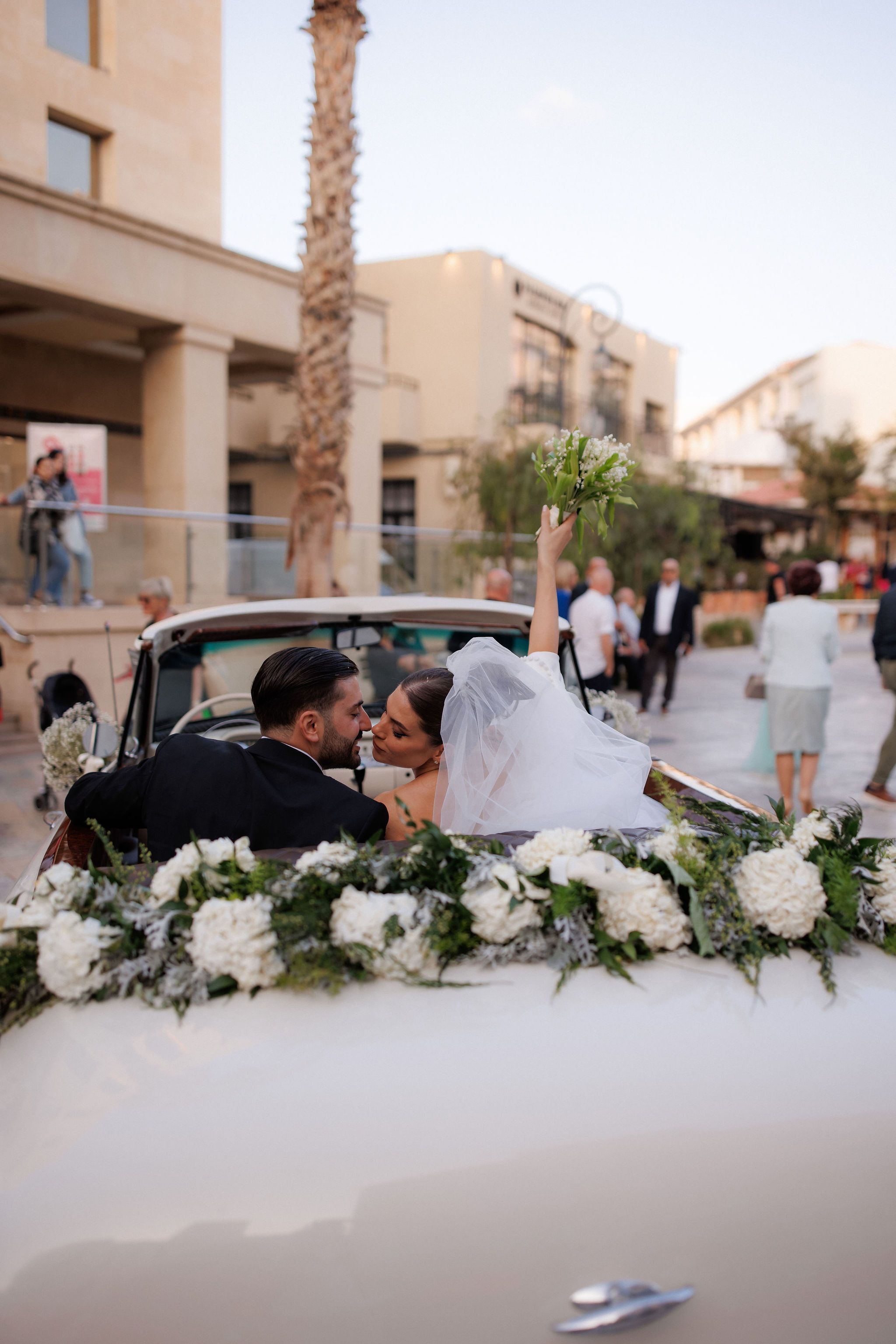 A bride in a white tulle veil and a dark-suited groom share a kiss in the back of a vintage white convertible, while she raises a small bouquet of white lily of the valley high in the air. The car is dressed with a garland of white hydrangeas and sprigs of silvery foliage, and guests stroll along a sunlit street behind them. These wedding car flowers were arranged by manfler in Greece.
