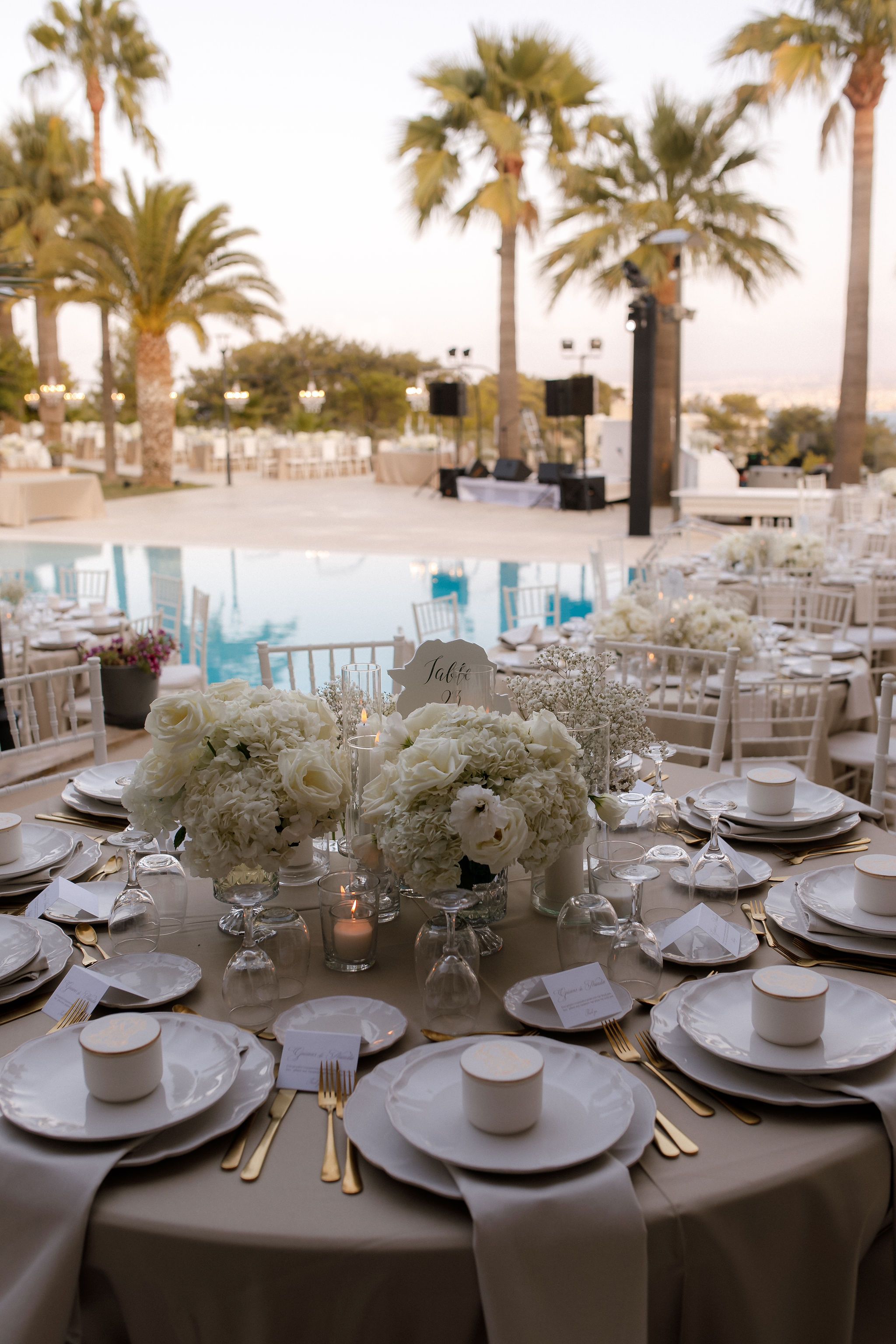 Round wedding tables in cream linen sit beside a pool at dusk, each centred with low arrangements of white roses and hydrangeas, with sprigs of gypsophila and small glass candle holders. A tall place card reading "Table" with a number stands among the flowers, and tall palm trees frame a pale sky above a staged dance floor at this wedding in Greece.