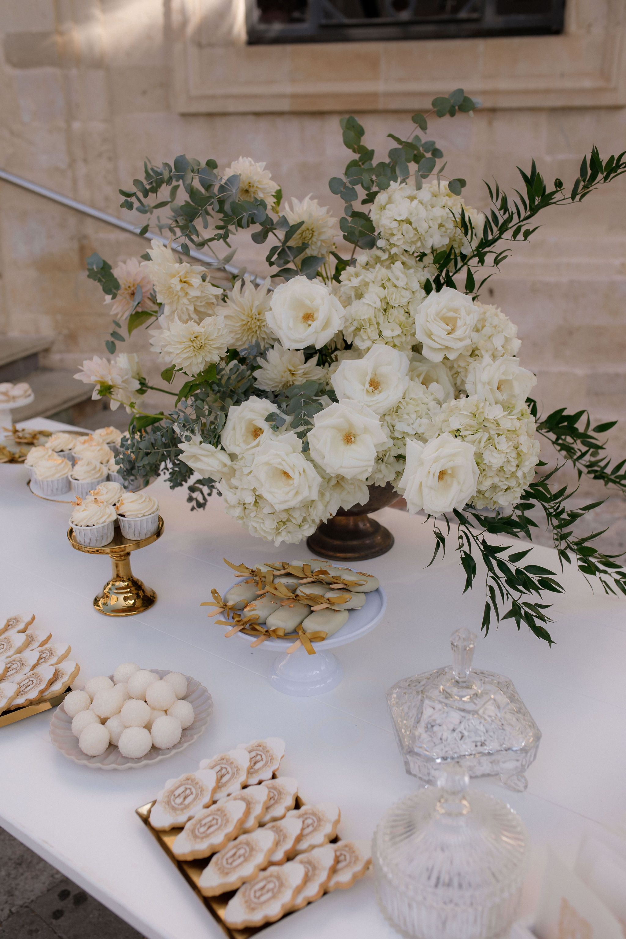 A tall, lush arrangement of cream garden roses, white hydrangeas and pale dahlias spills from a dark bronze pedestal urn, trailed with silvery eucalyptus and long green foliage. It sits on a white dessert table at a wedding in Greece, beside gold-rimmed cupcakes, glazed biscuits tied with gold ribbon and a cut-glass lidded dish, under soft natural light.