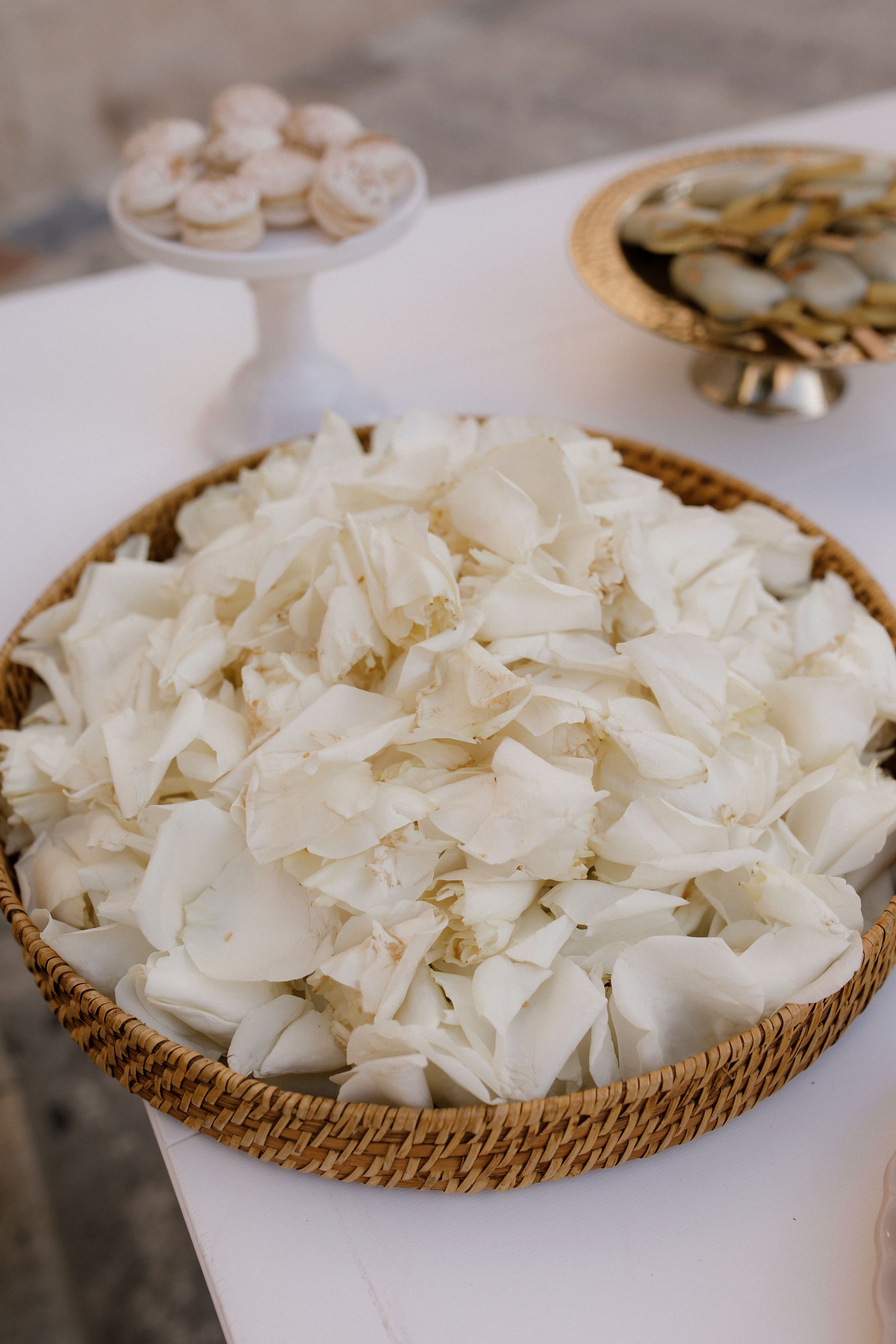 A woven wicker basket heaped with loose white rose petals, their creamy edges curling softly against one another, sits on a white tablecloth. Behind it, a gold-rimmed pedestal dish and a white candle or vase blur into the background, catching the soft daylight. These petals are wedding flowers from Manfler, ready to be scattered for a ceremony in Greece.