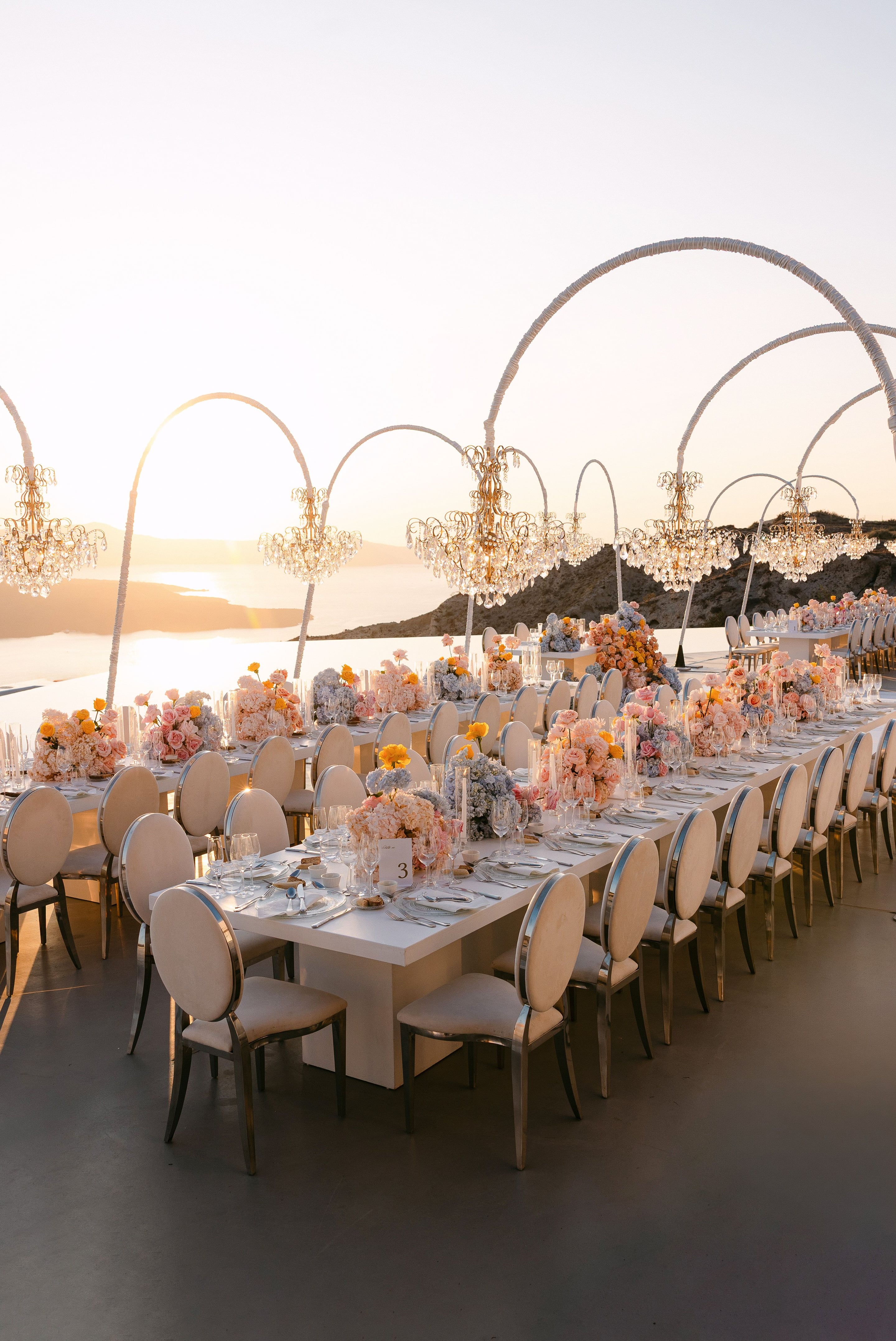 Long white banquet tables stretch along a clifftop terrace at Eros Santorini in Greece, set for a wedding dinner at sunset. Centrepieces of pale pink roses, peach hydrangeas, dusty blue hydrangeas and bright yellow ranunculus rise from the tablescape, beneath gold crystal chandeliers hung from white arched frames. Cream oval-backed chairs line the tables, and beyond the terrace the sea glows gold in the low evening light.
