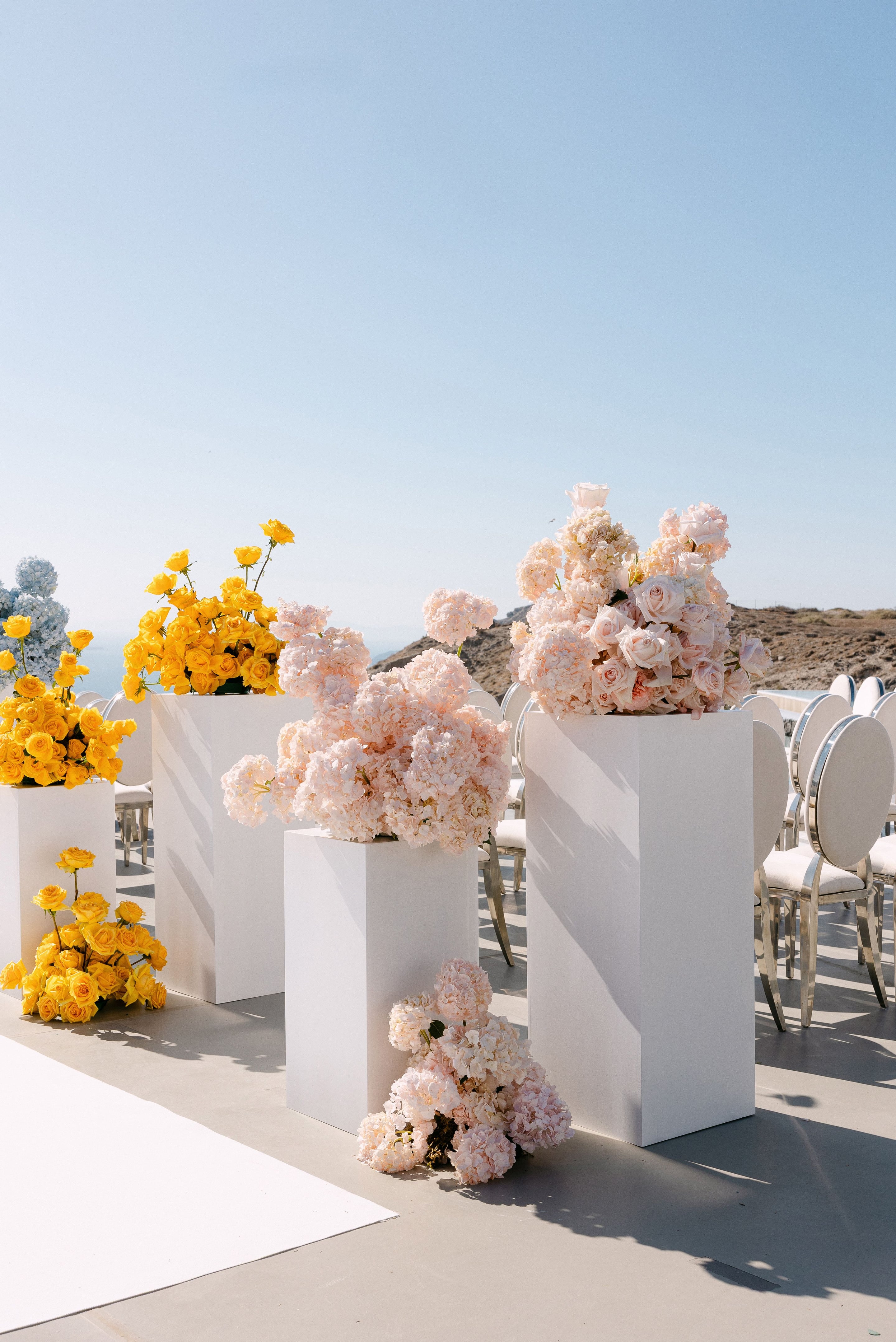 Tall white plinths line a sunlit terrace along a wedding aisle, topped and framed with blush hydrangeas and pale pink garden roses on the right, and cascades of yellow roses on the left. A white runner stretches across the grey floor beside rows of silver-framed, cream-cushioned chairs, with a dry hillside and the sea beyond. This is a wedding ceremony setting at Eros Santorini in Greece.