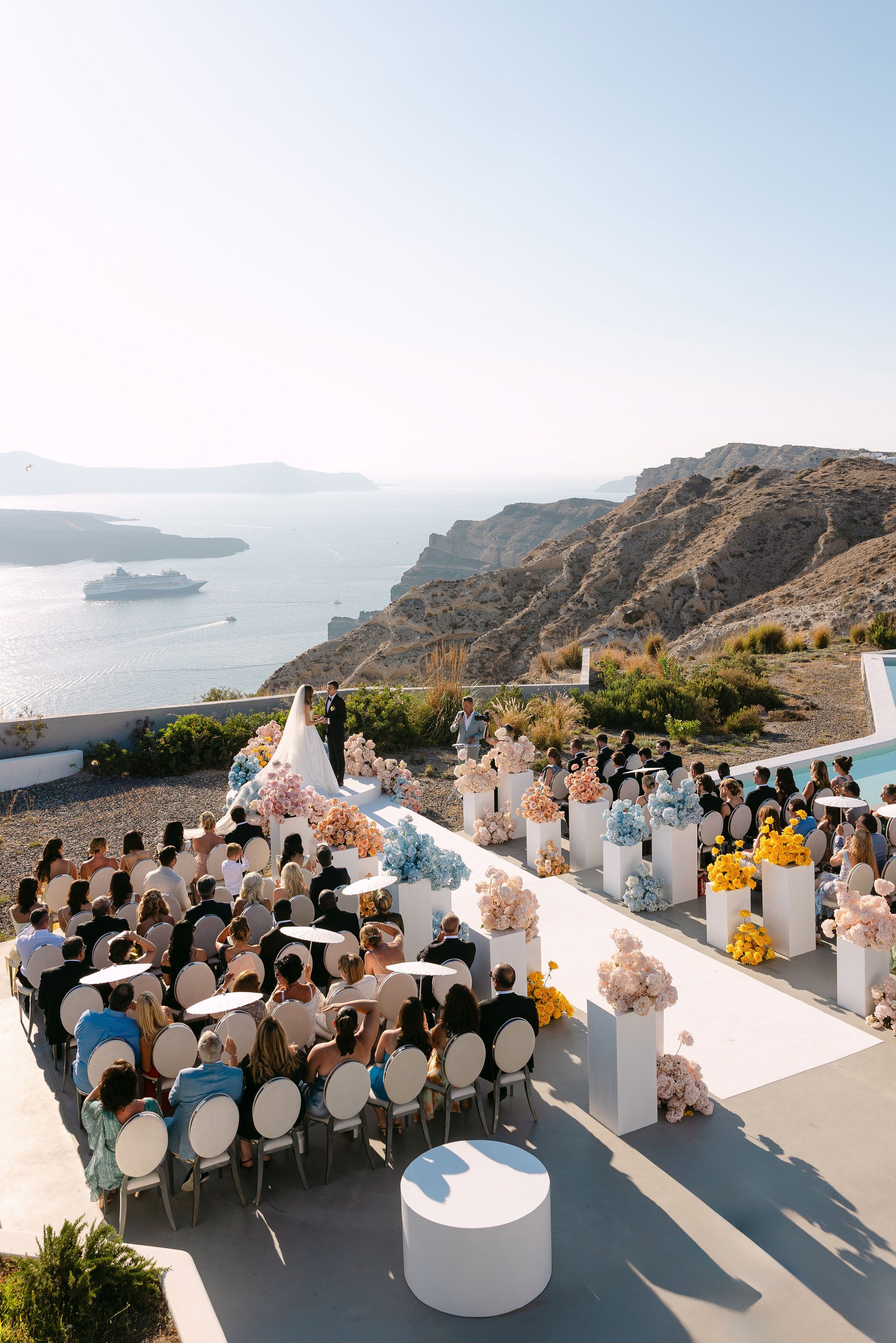 A wedding ceremony on a clifftop terrace at Eros Santorini, Greece, seen from above, with guests seated on white oval chairs facing a white aisle lined with tall white plinths of pink, peach, blue and yellow hydrangeas and roses. The bride in a long veil and the groom in a dark suit stand at a small raised altar, with the sea and a cruise ship in the distance and dry hillside scrub around the terrace.