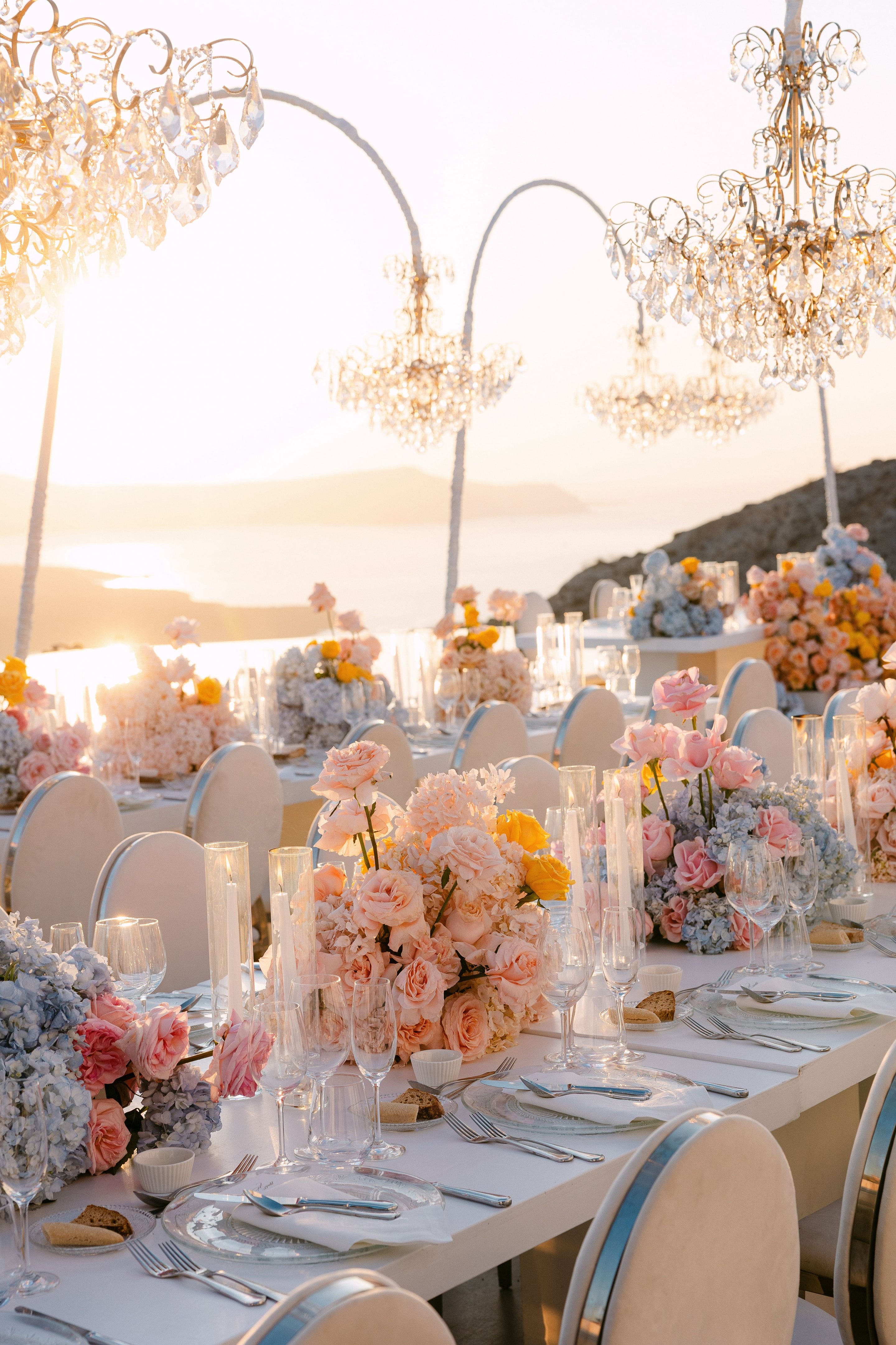 A long wedding dinner table at sunset, set on a terrace at Eros Santorini in Greece, with the caldera sea glowing gold behind it. Low centrepieces of blush garden roses, pale peach hydrangeas and sprigs of yellow roses spill over the white cloth, interspersed with powder-blue hydrangeas. Tall taper candles stand in glass cylinders between clear glasses and cream chairs, and the warm light gives the whole scene a soft, romantic glow.