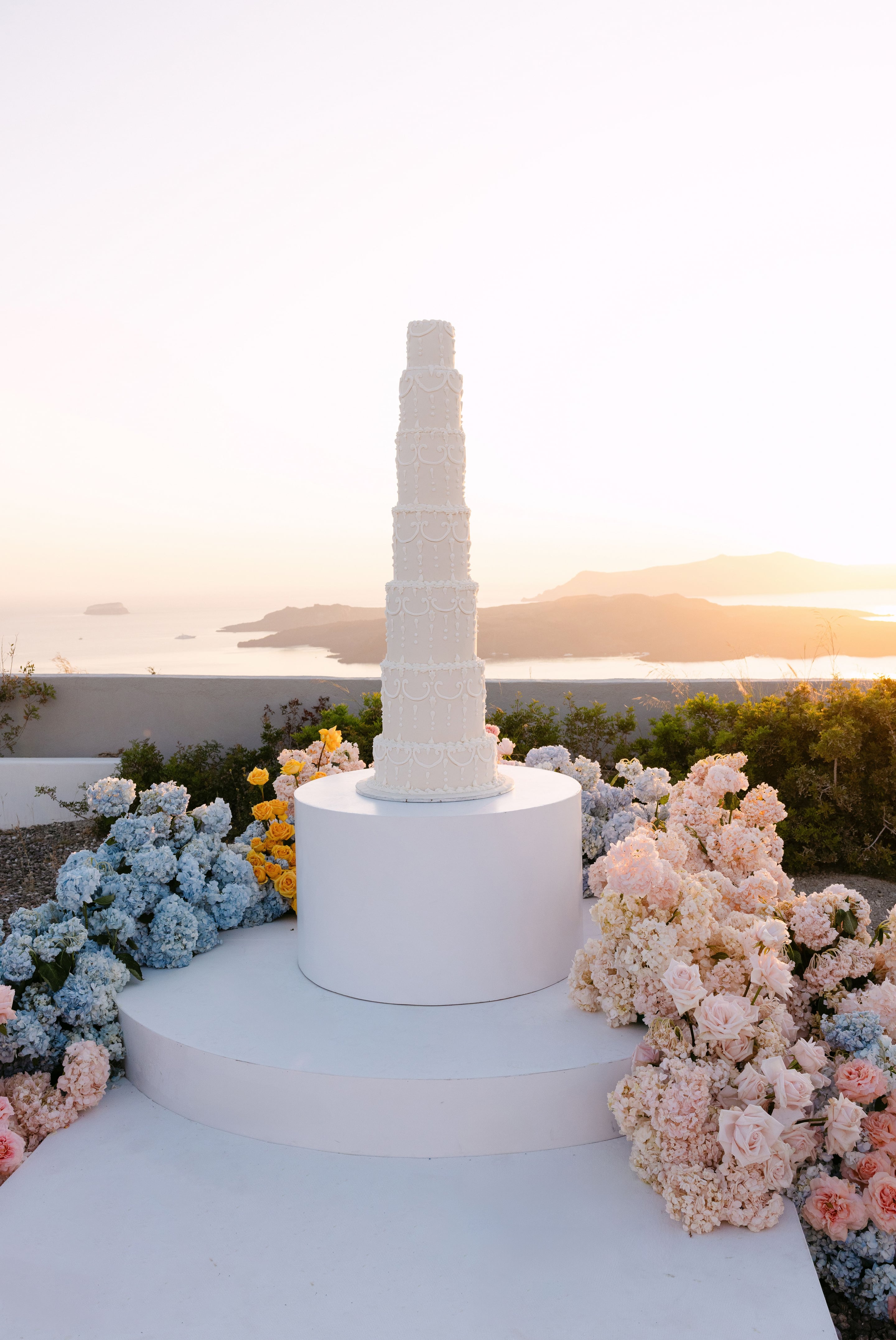 A tall, tiered white wedding cake, piped with delicate scrollwork and beading, stands on a white circular pedestal at Eros Santorini in Greece, framed by drifts of pale blue hydrangeas, blush roses and cream hydrangeas, with small yellow roses tucked in. The warm, golden sunset light softens the scene against a hazy view of the Aegean and distant islands.