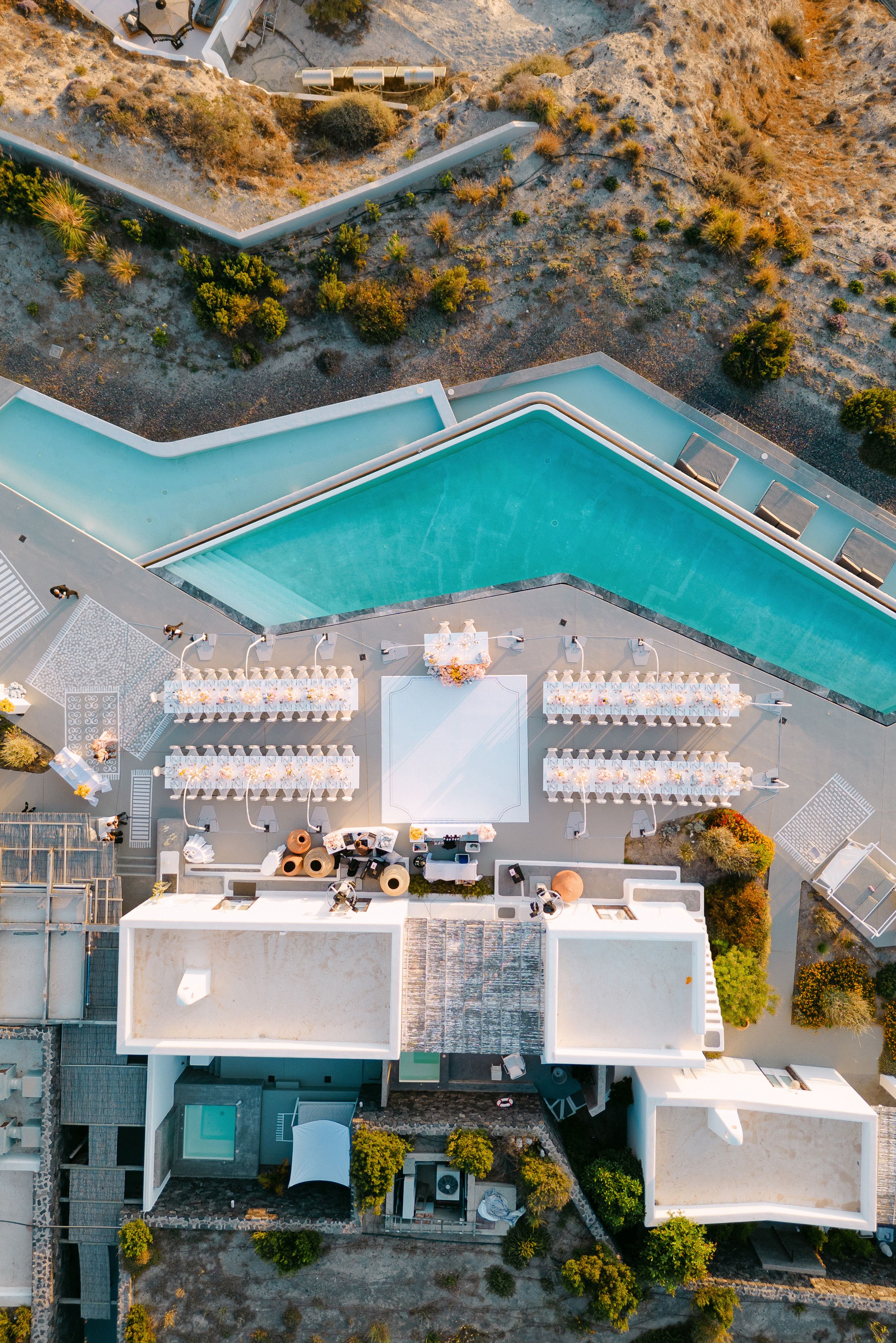 An overhead aerial view of a wedding reception at Eros Santorini in Greece, showing four long banquet tables dressed in white linen and lined with low arrangements of cream and pale peach blooms, likely roses and orchids. A large white dance floor sits at the centre beside a floral centrepiece, all beside a turquoise infinity pool and white-washed villa rooftops, with a sunlit terrace and a strip of golden-orange shrubs along the edge.