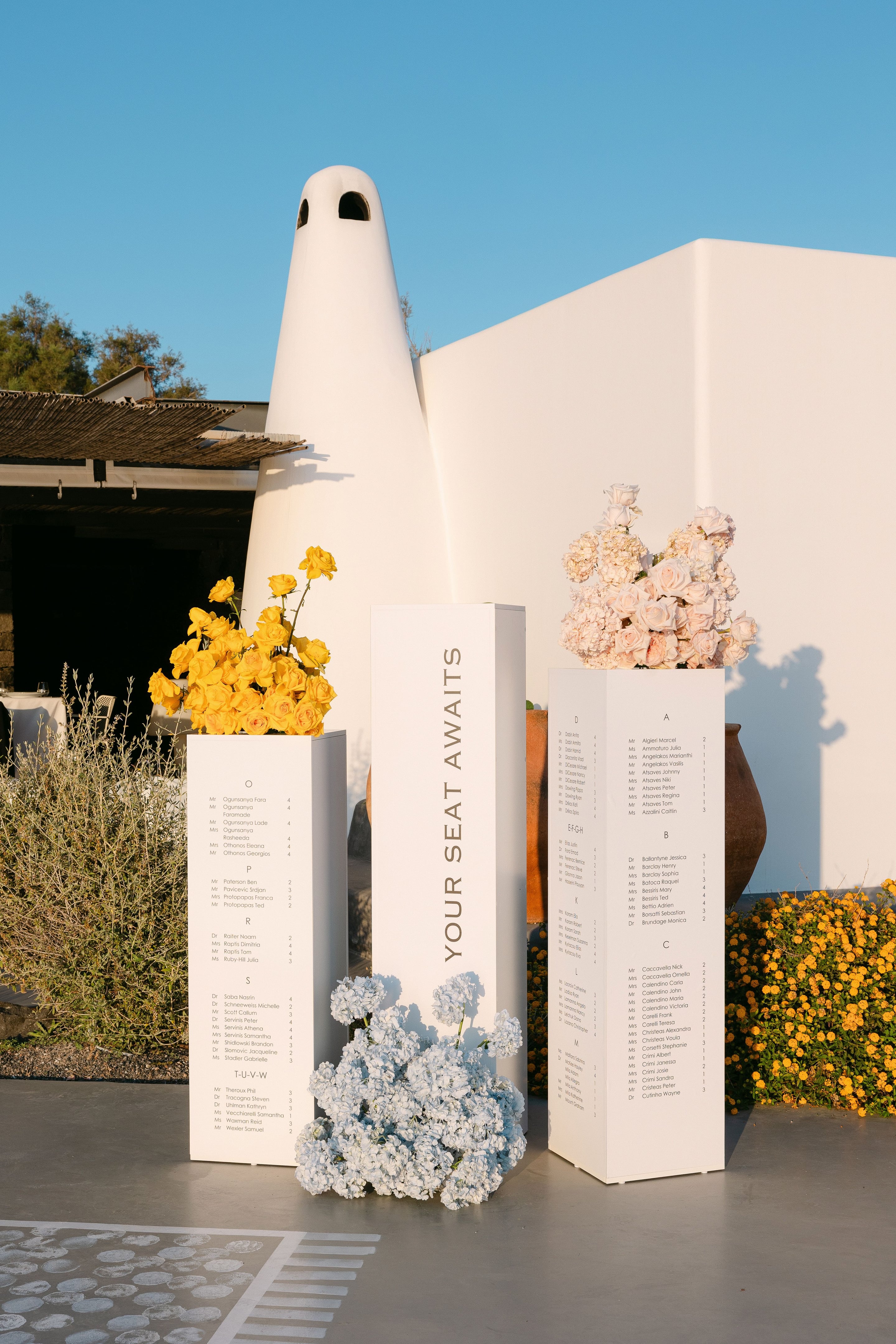 Three tall white plinths stand on a grey terrace at Eros Santorini in Greece, each printed with alphabetised wedding seating names, with the central one reading "Your seat awaits" in grey letters. Bright yellow roses spill from the left plinth, pale cream roses and hydrangea from the right, and a low cascade of pale blue hydrangeas sits at the base of the middle one, all against a whitewashed wall in warm evening light.