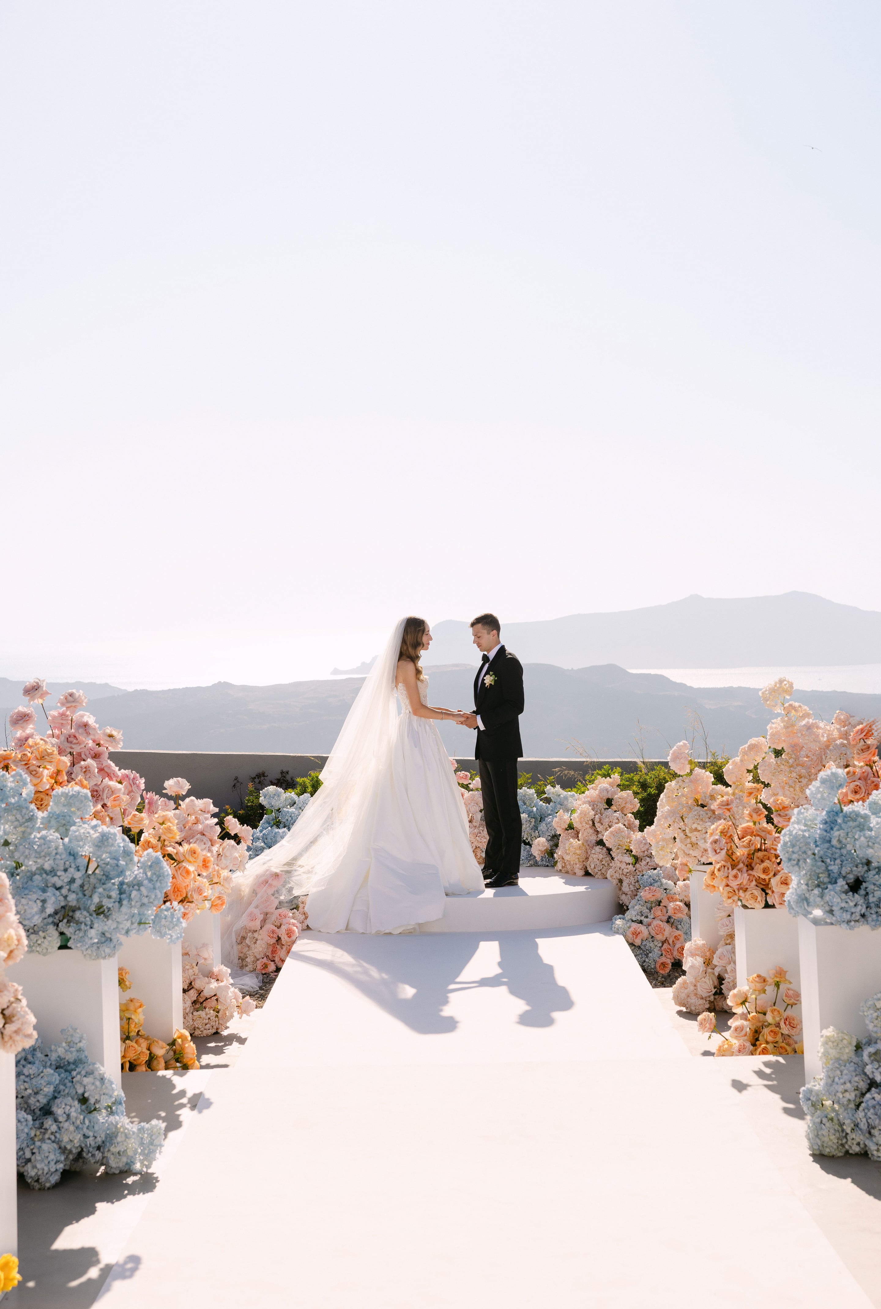 A bride in a white ball gown with a long veil and a groom in a black tuxedo face each other, holding hands on a raised white platform at the end of a white aisle. Both sides of the aisle are lined with towering pale blue hydrangeas, peach garden roses and cream blooms in white plinths, all set against a hazy view of distant hills and sea at Eros Santorini, a wedding venue in Santorini, Greece.