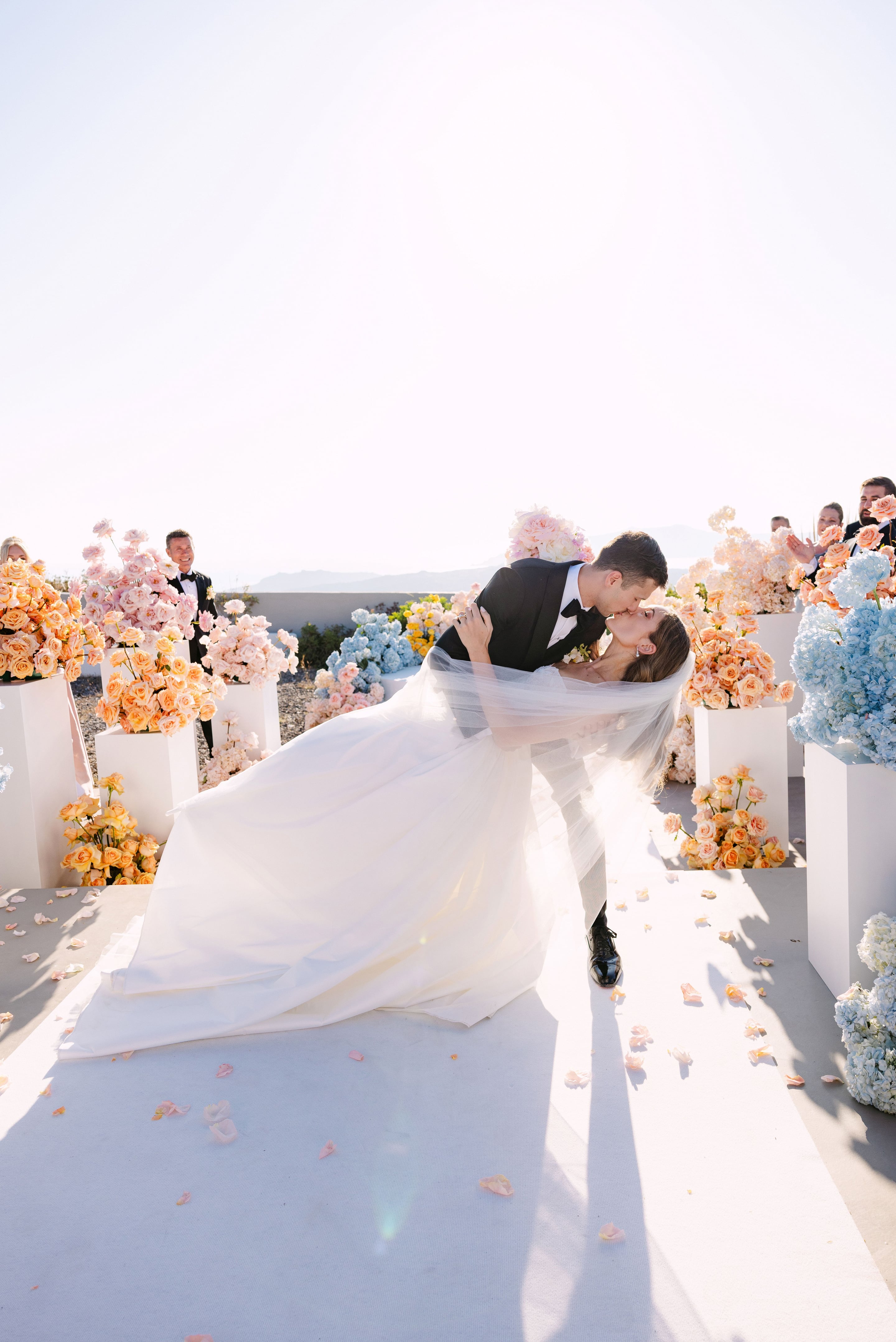 A groom in a black tuxedo dips his bride in a flowing white gown and sheer veil for a kiss on a sunlit terrace, with pale hazy sky behind them. Along the white aisle stand tall white plinths overflowing with peach garden roses, pale pink blooms, and sky-blue hydrangeas, while rose petals scatter the path and guests applaud on the right. This is a wedding flower display at Eros Santorini in Greece.