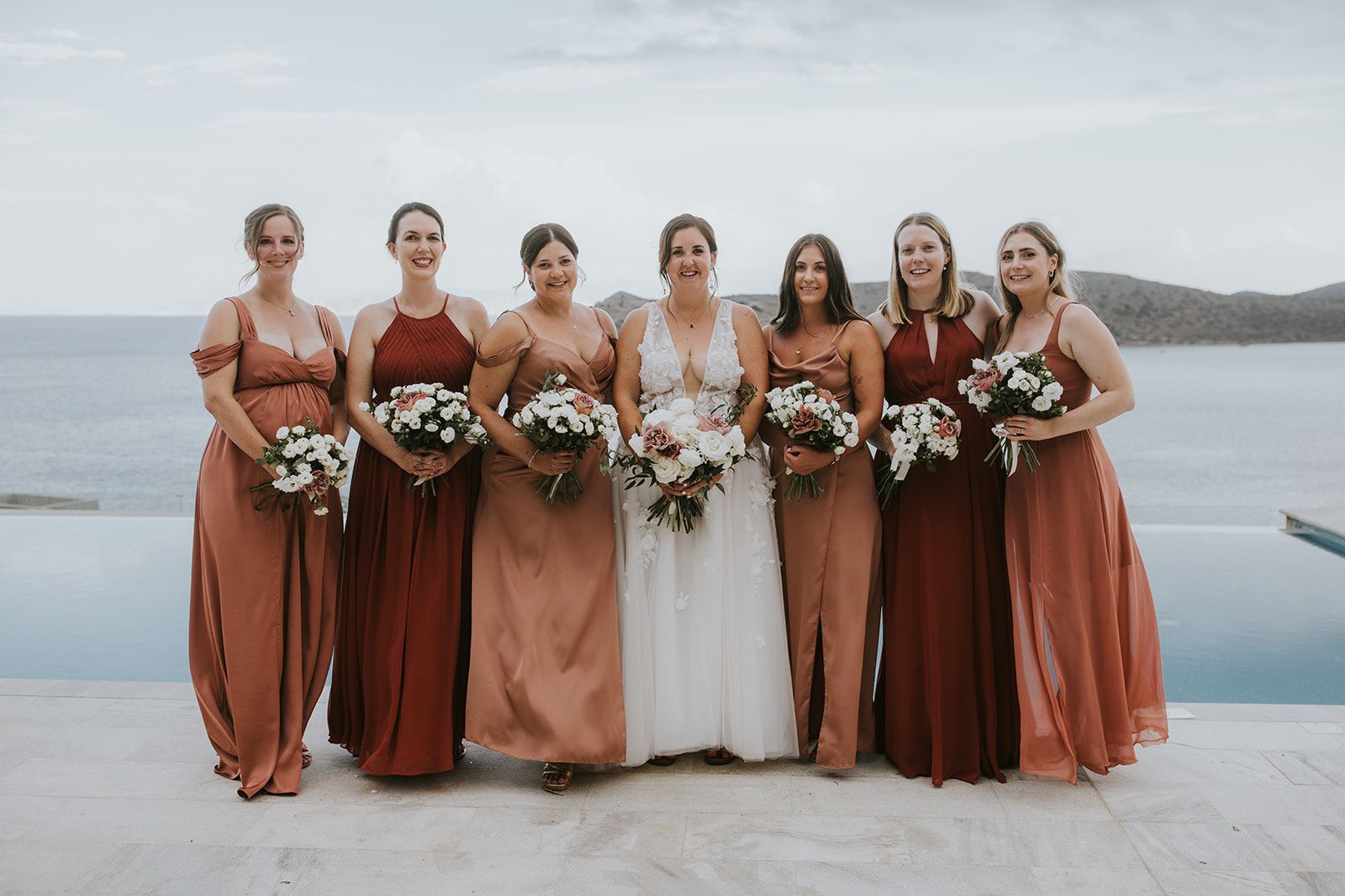 Seven women stand in a row on a pale stone terrace beside an infinity pool, with grey sea and a rocky headland behind them under an overcast sky. The bride at the centre wears a white lace gown with a deep V neckline and holds a full bouquet of white roses, blush garden roses and greenery. Her six bridesmaids, in flowing terracotta, rust and dusty-rose gowns, each carry smaller round bouquets of white blooms with dusty-pink accents. This wedding flower display is from Villa Gina in Greece.