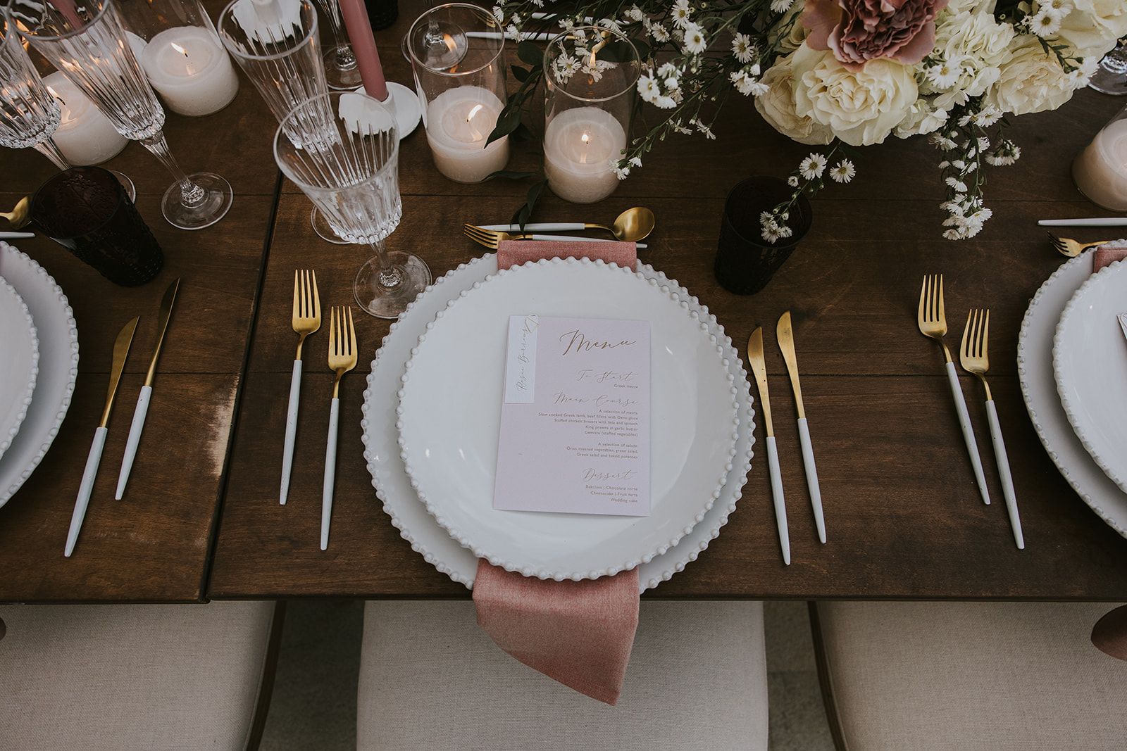 A wedding table setting at Villa Gina in Greece, seen from above on a dark wooden table. A place setting of beaded white plates sits on a dusty-pink velvet napkin, with gold and white-handled cutlery and a blush menu card in gold script. Behind it, cream roses, blush garden roses, and sprigs of white daisies with dark greenery rise from a centrepiece beside glowing pillar candles in glass holders.