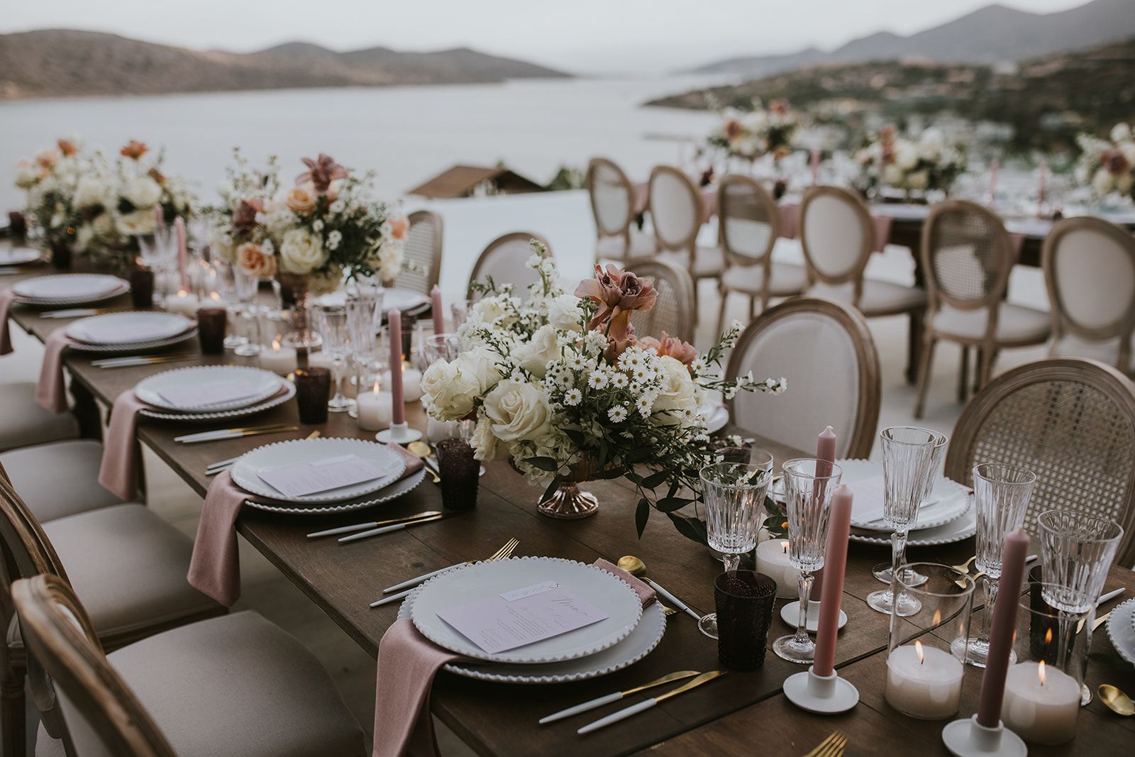 A long wooden wedding table set on a terrace overlooking the sea at Villa Gina in Greece, with a centrepiece of white roses, white daisies and dusky pink blooms in a low urn. Blush linen napkins, pale scalloped plates, gold cutlery and pink taper candles run down the table, with more flower arrangements and cane-backed chairs stretching into the soft evening light.
