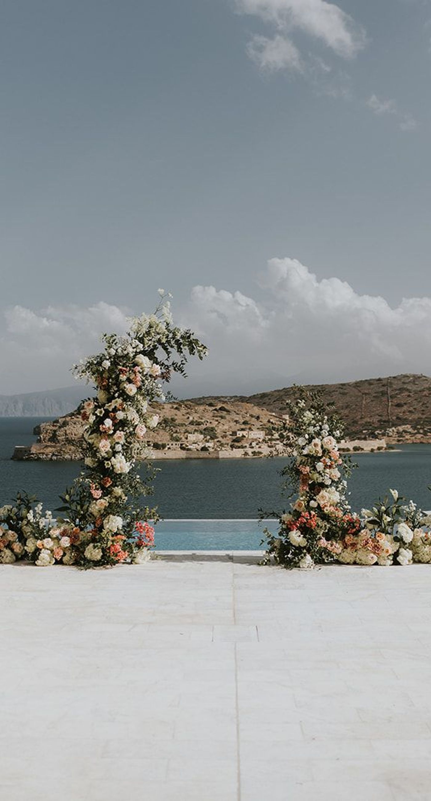 Nina& Samouel - In a beautiful symphony of nature and romance, this scene captures the essence of a perfect coastal celebration. The gentle breeze carries sweet floral whispers as the couple walks amidst a cascade of pastel petals, their smiles reflecting joy and love. The soft blues of the ocean and sky provide a serene backdrop, harmonizing with the lush greenery and delicate whites and pinks of the blooms. This composition encapsulates an idyllic, nature-infused elegance, showcasing the artistry and thoughtful design of the florist's craft. - Photo 2