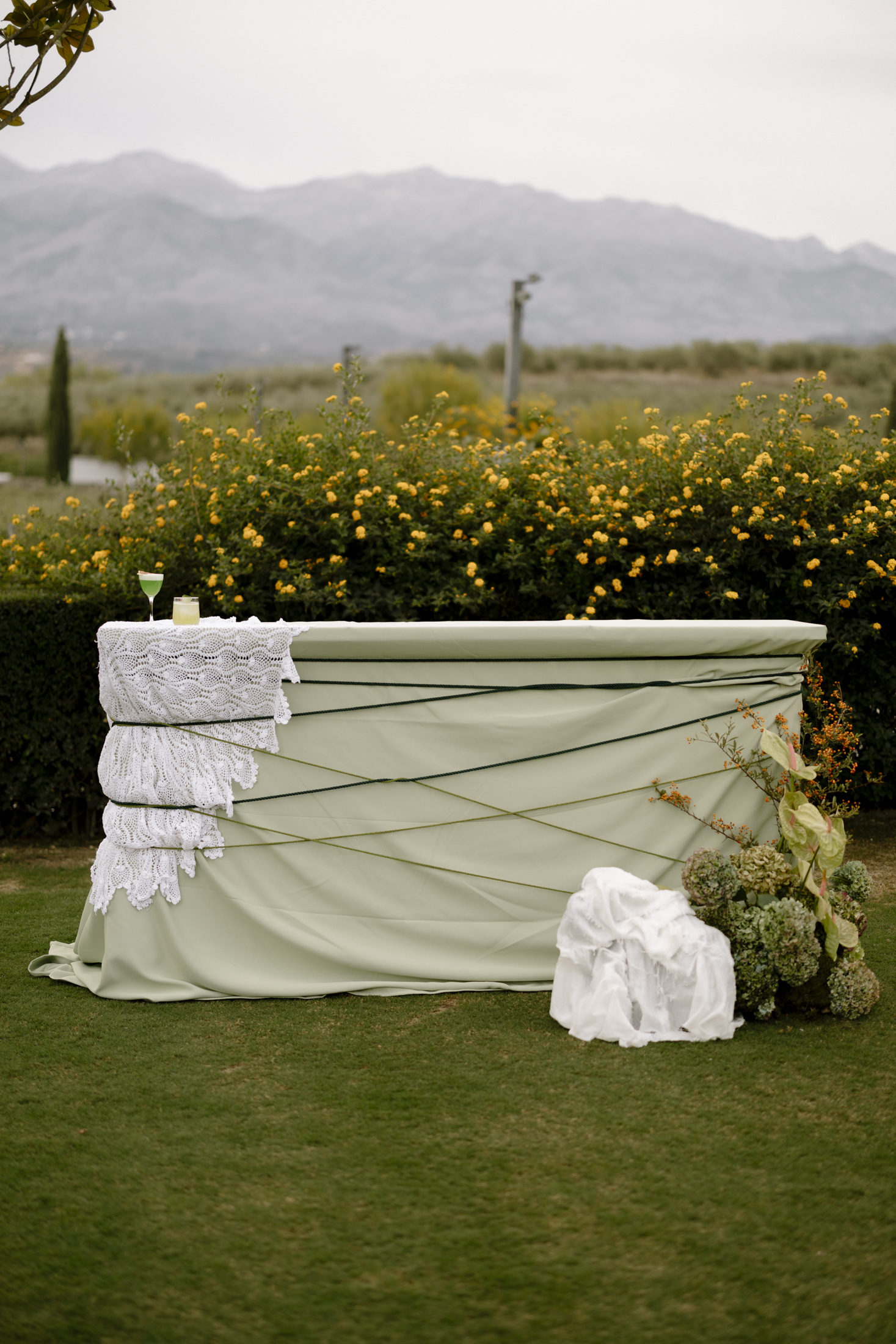 A sage-green draped bar stands on lawn in front of a dense hedge of small yellow flowers at Roxani Estate in Greece, styled for a wedding. Its front is wrapped in dark green cords, and the top is layered with stacked white crocheted lace runners holding two cocktail glasses, one with a pale green drink. At the right, a loose cluster of green hydrangeas, pale green anthurium and sprigs of orange berries spills beside a crumpled white cloth.