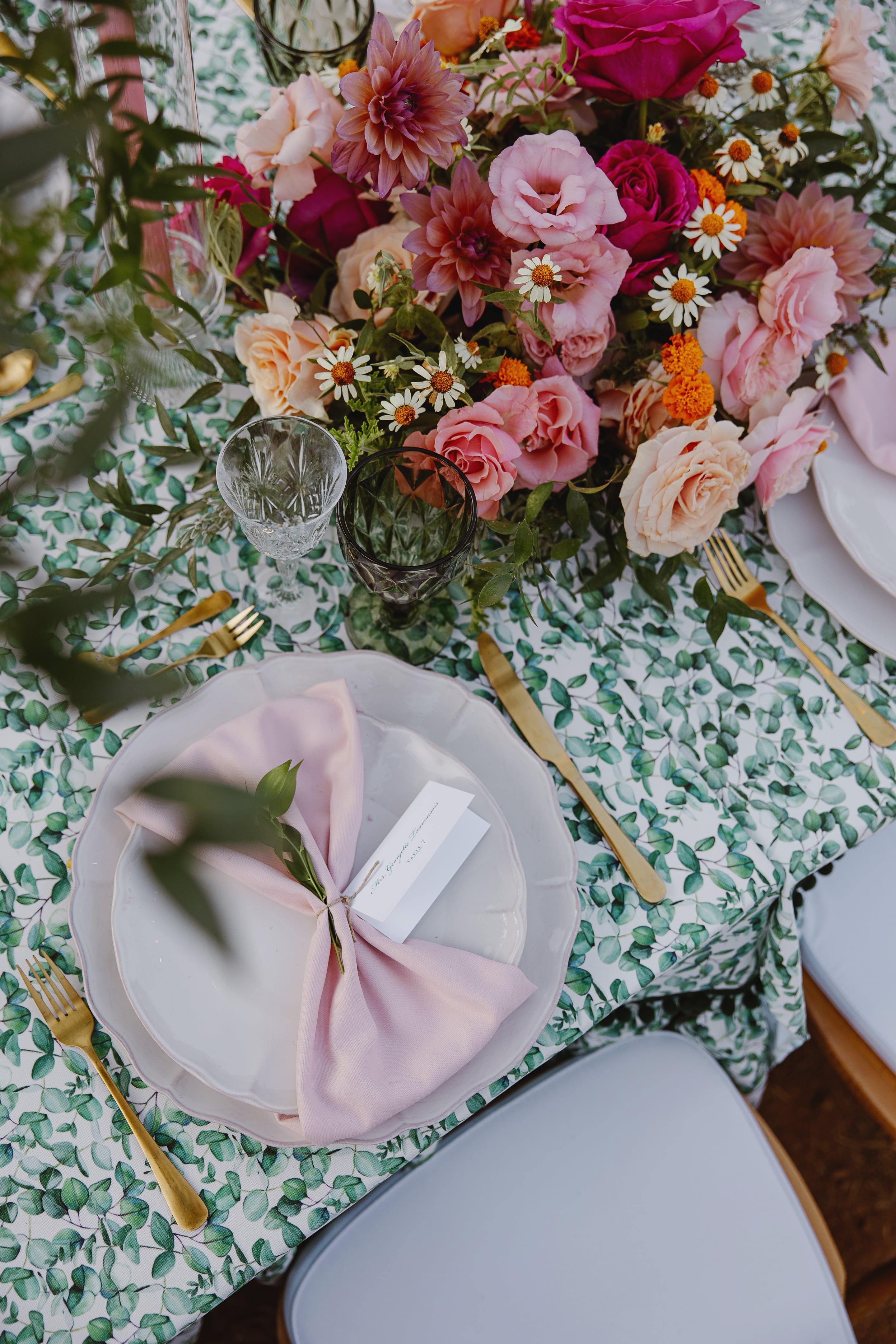 A top-down view of a wedding table set on a leaf-patterned cloth, with a lush centrepiece of pink garden roses, peach roses, dahlias, white daisies and orange marigolds. Gold cutlery, crystal and smoky green glasses, and a blush napkin on a white plate with a name card frame the arrangement, in Greece.