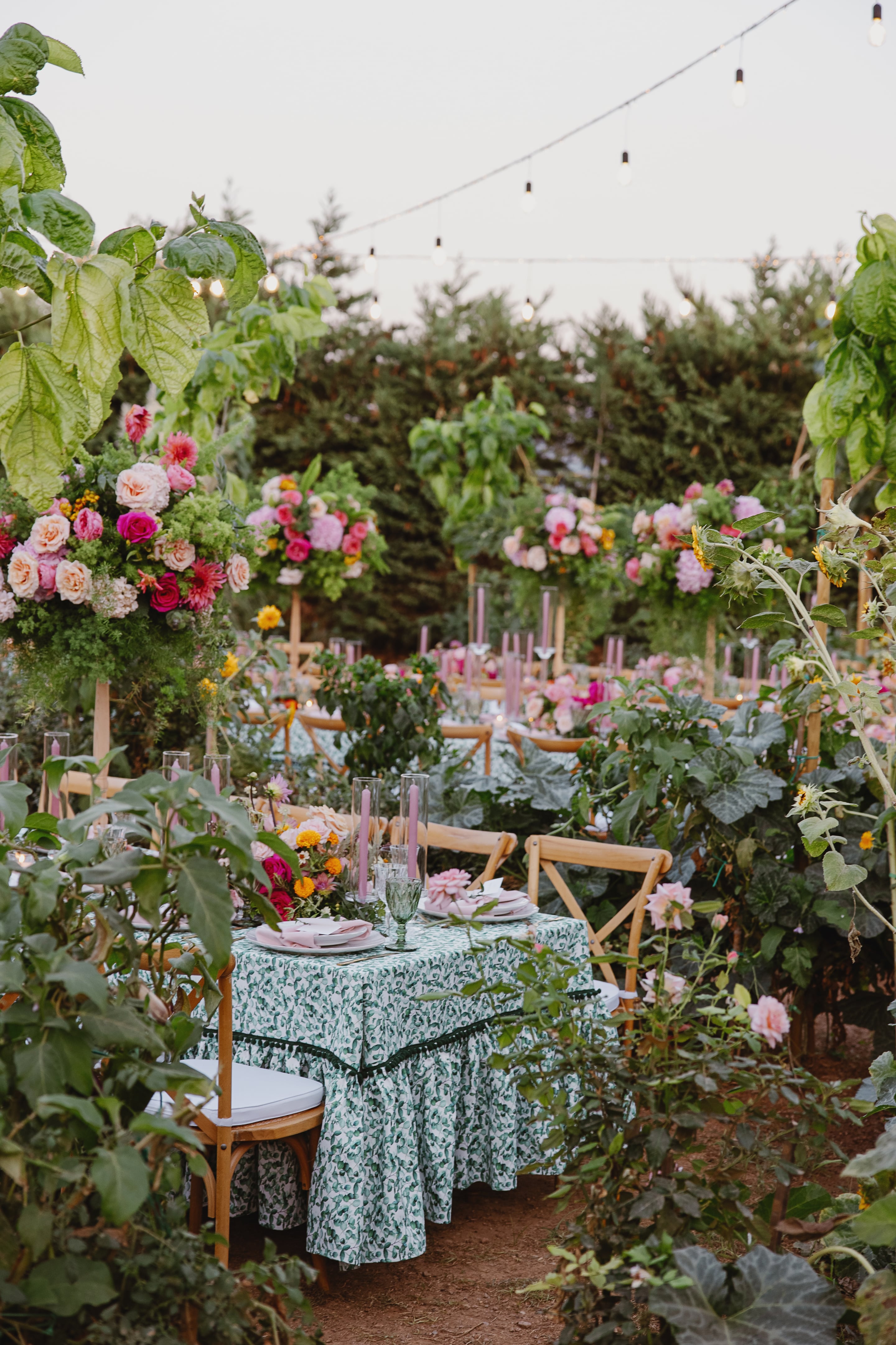 A wedding table setting in a lush garden in Greece, with a green and white patterned cloth, pink plates and lilac candles in glass hurricanes. Behind it, tall floral arrangements of pink roses, peonies and dahlias on wooden stands rise among leafy green plants and sunflowers, with rows of cross-back chairs stretching into the greenery.