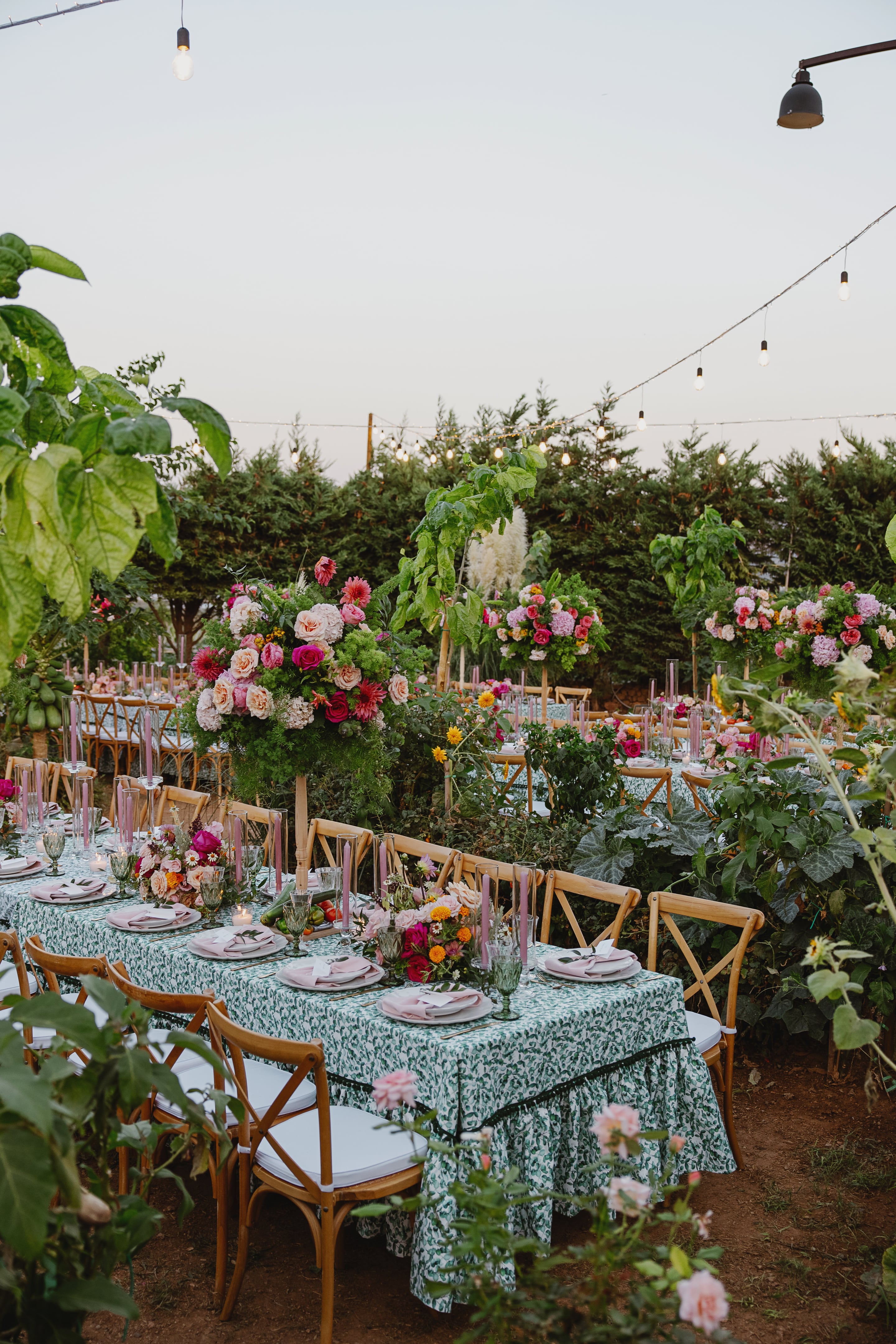 Long wedding tables in an open garden in Greece, dressed in a green leaf-print cloth with a ruffled hem and set with pink napkins and tall pink taper candles. Tall floral centrepieces of garden roses, pink dahlias, hydrangeas and peach blooms rise above the table settings, with smaller arrangements of marigolds and roses along the runners. Cross-back wooden chairs with white cushions line the tables, framed by lush green foliage and string lights overhead in the soft evening light.