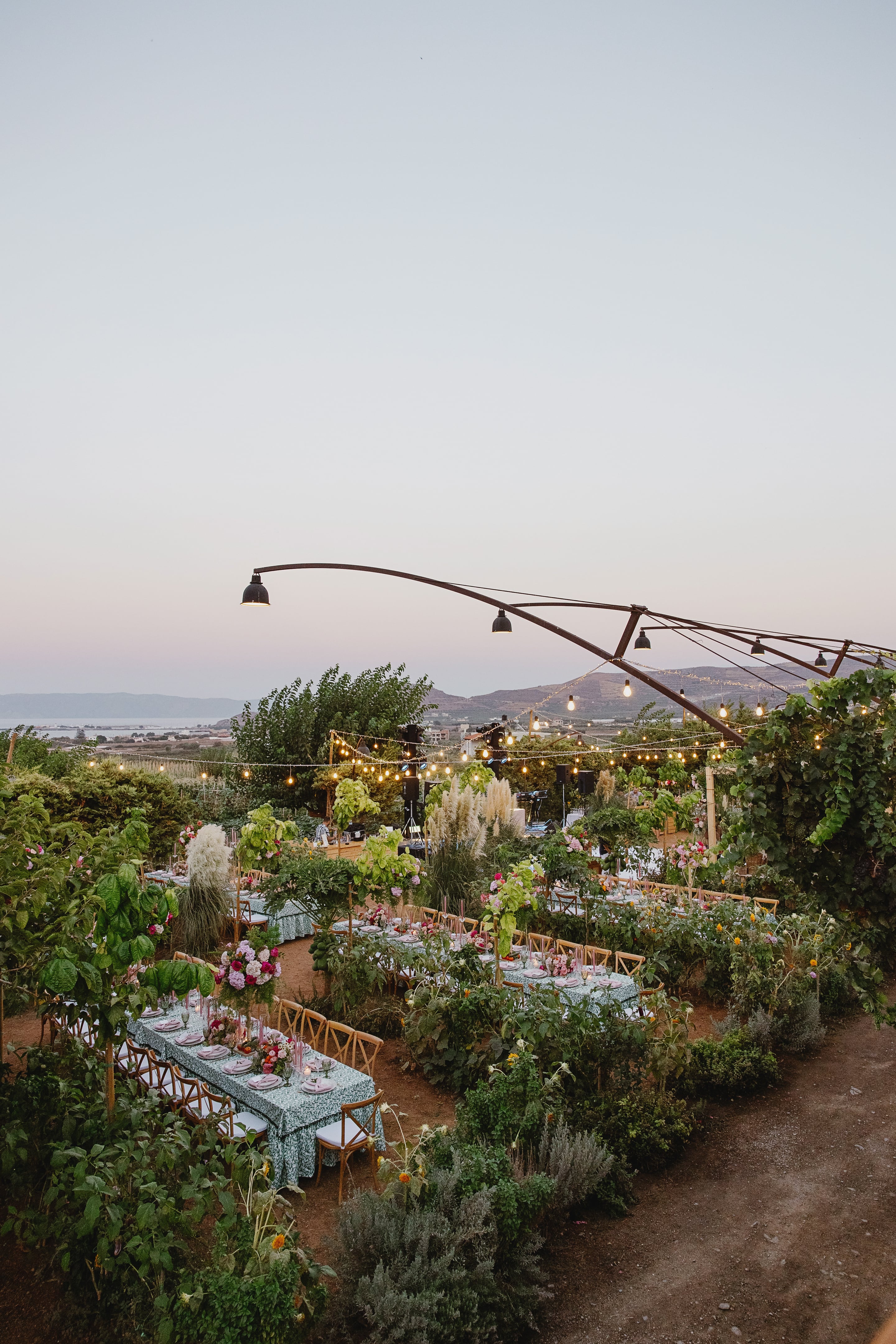 A wide dusk view of long wedding banquet tables set among lush garden greenery, with pink and white flower centrepieces, pampas grass plumes and string lights overhead, on a terrace in Greece. Cross-back chairs line the tables in rows beneath a curved metal frame, with soft pale-lilac sky and distant hills behind.