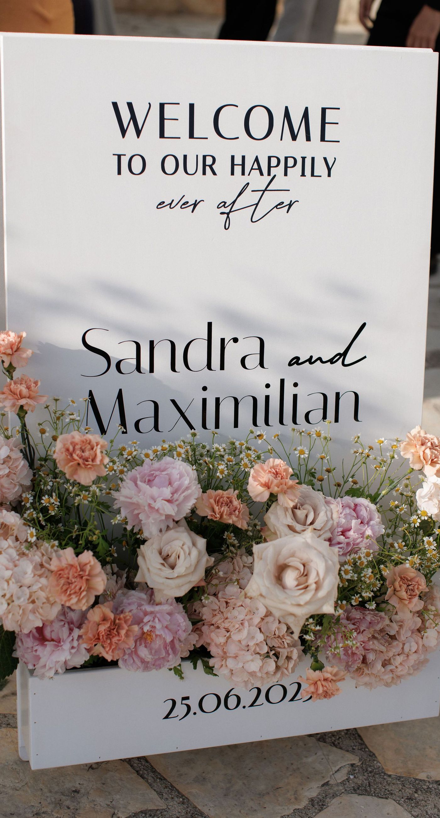 Sandra & Maximilian - In a serene garden setting, a couple stands embraced, surrounded by a breathtaking floral display. The bride's off-the-shoulder lace gown complements the groom's classic black tuxedo, both exuding elegance amid the lush, natural backdrop. Pastel blooms of blush and cream, expertly arranged, create an arch that seamlessly blends with the verdant landscape. Soft pinks and whites, interspersed with touches of greenery, add a delicate yet sophisticated touch. Rolling hills and distant mountains frame the scene, enhancing the timeless beauty and tranquility of this picturesque moment. - Photo 3