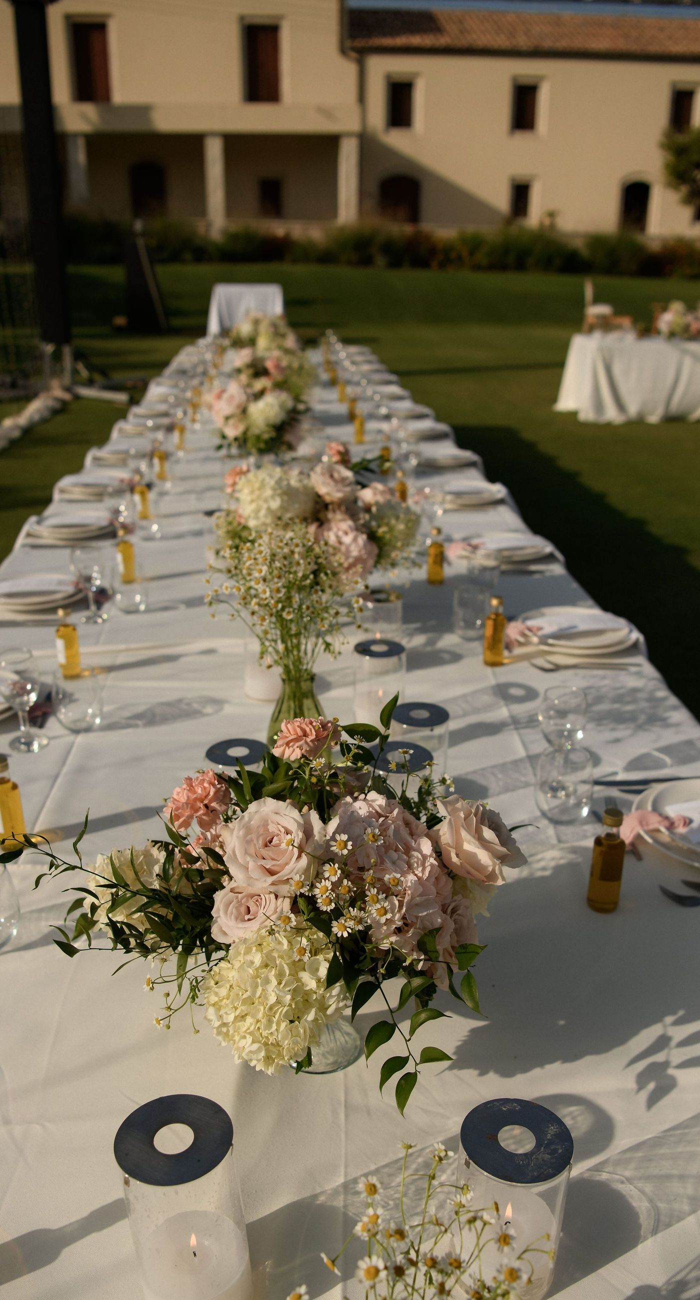 Sandra & Maximilian - In a serene garden setting, a couple stands embraced, surrounded by a breathtaking floral display. The bride's off-the-shoulder lace gown complements the groom's classic black tuxedo, both exuding elegance amid the lush, natural backdrop. Pastel blooms of blush and cream, expertly arranged, create an arch that seamlessly blends with the verdant landscape. Soft pinks and whites, interspersed with touches of greenery, add a delicate yet sophisticated touch. Rolling hills and distant mountains frame the scene, enhancing the timeless beauty and tranquility of this picturesque moment. - Photo 11