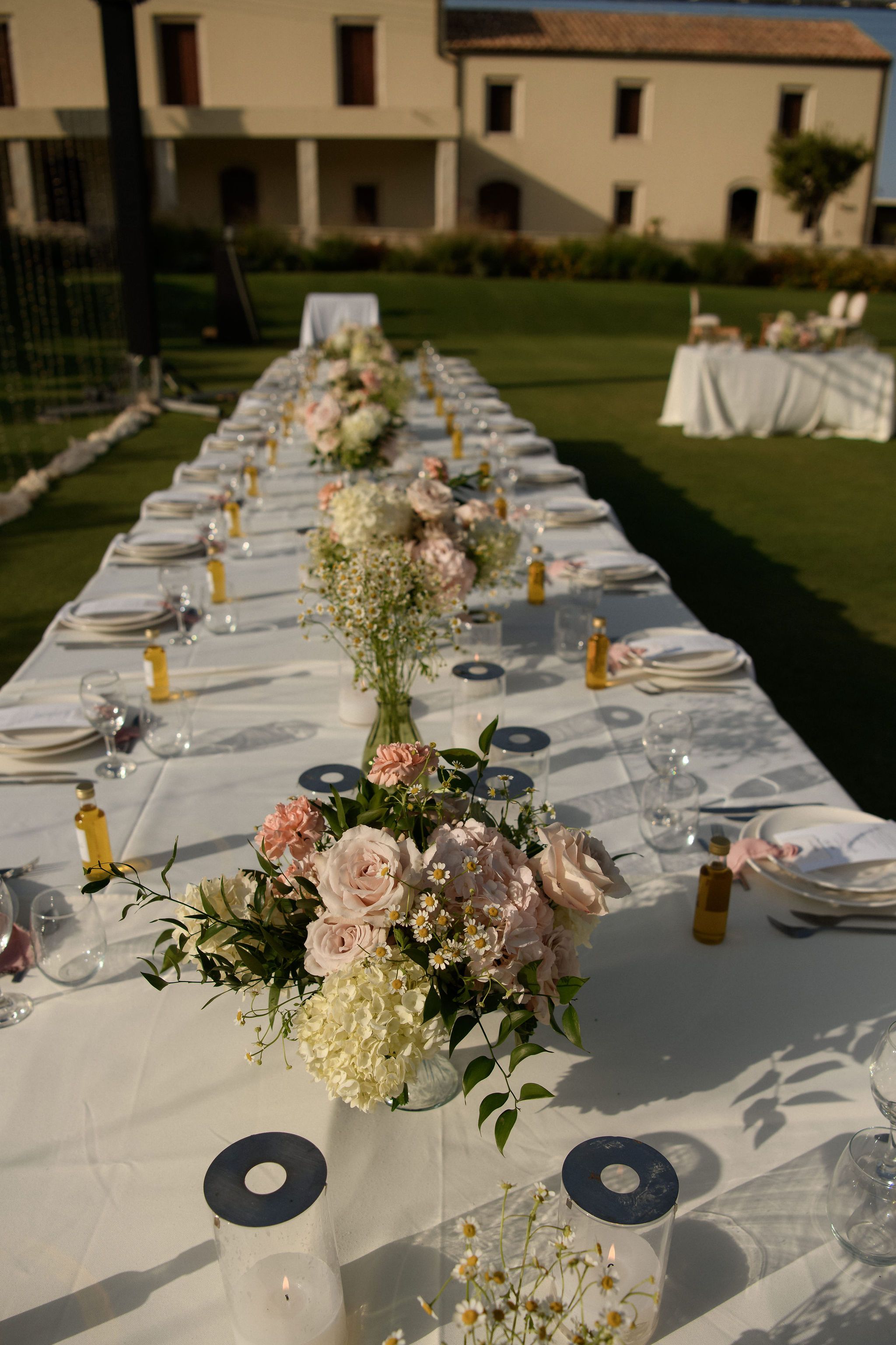 A long white-clothed wedding table stretches across a sunlit lawn at the Roxani estate in Greece, lined with white plates, glasses and small bottles of golden olive oil. Low arrangements of blush garden roses, peach carnations, white hydrangeas and tiny chamomile daisies run down its length, with a glass vase of wildflowers in the centre. In the soft background, another table dressed in white stands beneath the estate's stone building.