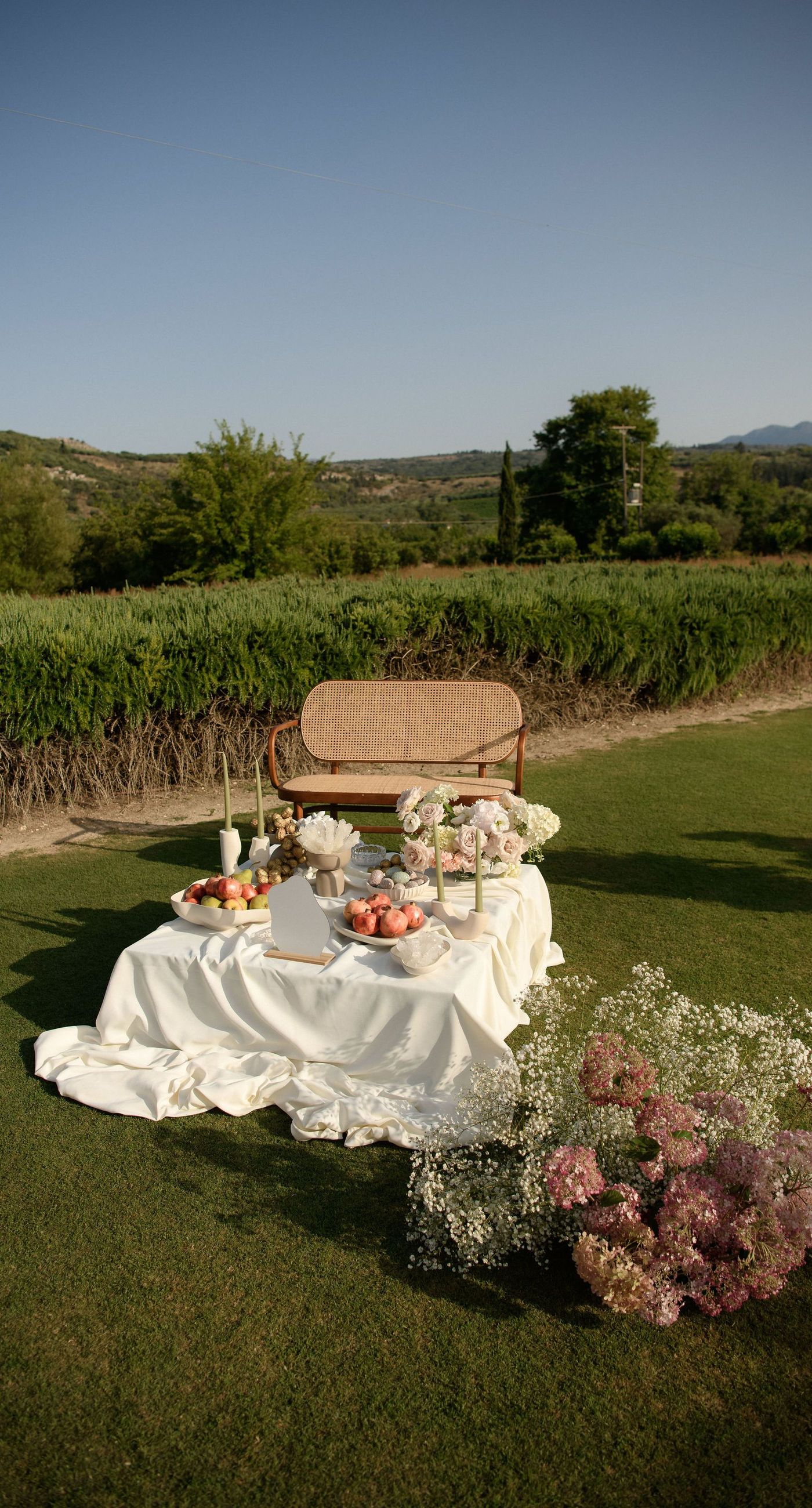 Sandra & Maximilian - In a serene garden setting, a couple stands embraced, surrounded by a breathtaking floral display. The bride's off-the-shoulder lace gown complements the groom's classic black tuxedo, both exuding elegance amid the lush, natural backdrop. Pastel blooms of blush and cream, expertly arranged, create an arch that seamlessly blends with the verdant landscape. Soft pinks and whites, interspersed with touches of greenery, add a delicate yet sophisticated touch. Rolling hills and distant mountains frame the scene, enhancing the timeless beauty and tranquility of this picturesque moment. - Photo 9