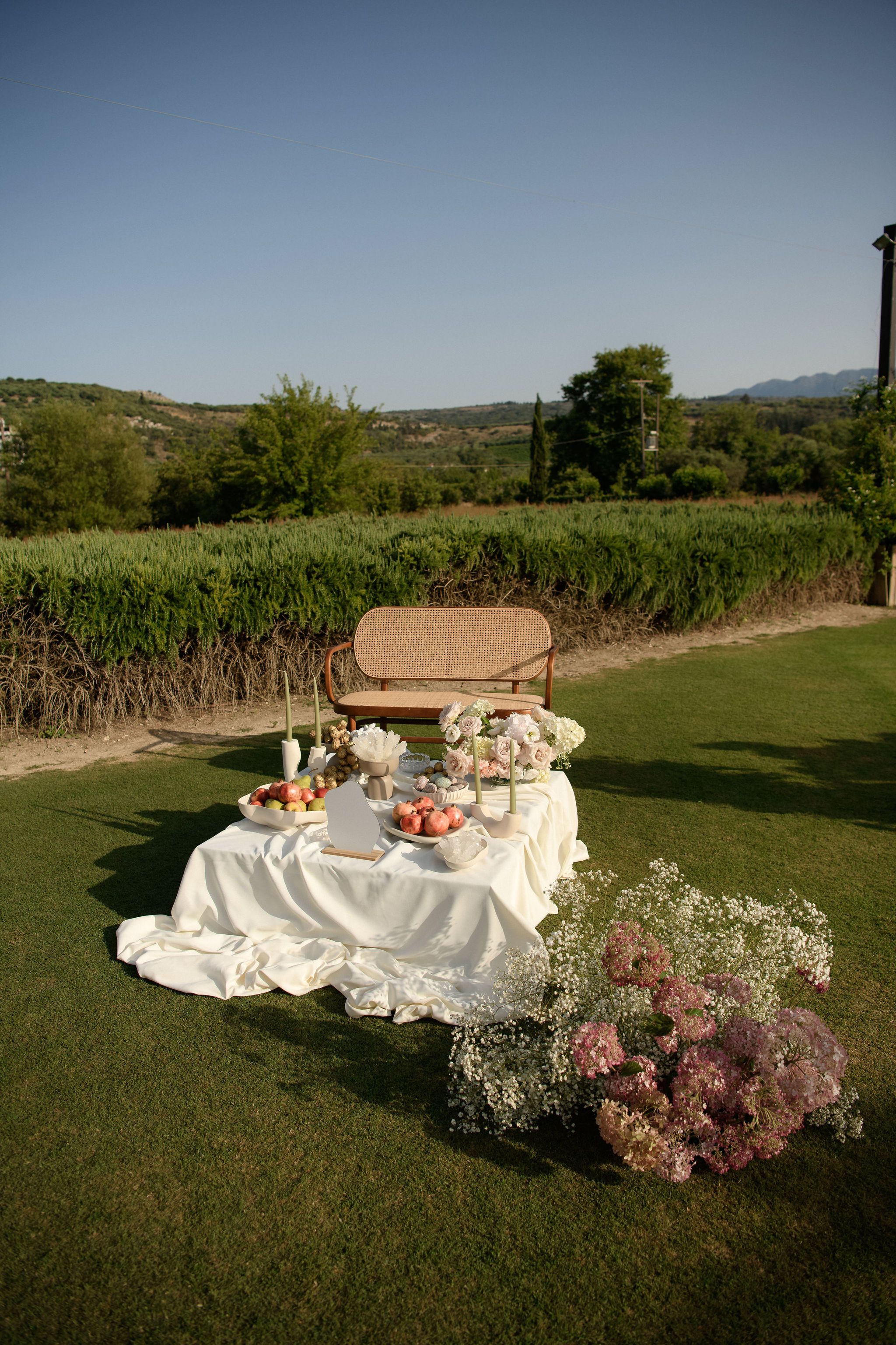 A romantic picnic set on a lawn at the Roxani estate in Greece, where a long white linen cloth spills across the grass in soft folds. Behind it a cane-backed wooden bench sits in front of a tall green hedge, and a bowl of pomegranates, apples and grapes rests on the table beside taper candles in pale sage. In the foreground, a loose cascade of baby's breath and blush-pink hydrangeas tumbles across the grass, all bathed in warm golden evening light, these being wedding flowers for Sandra and Maximilian.