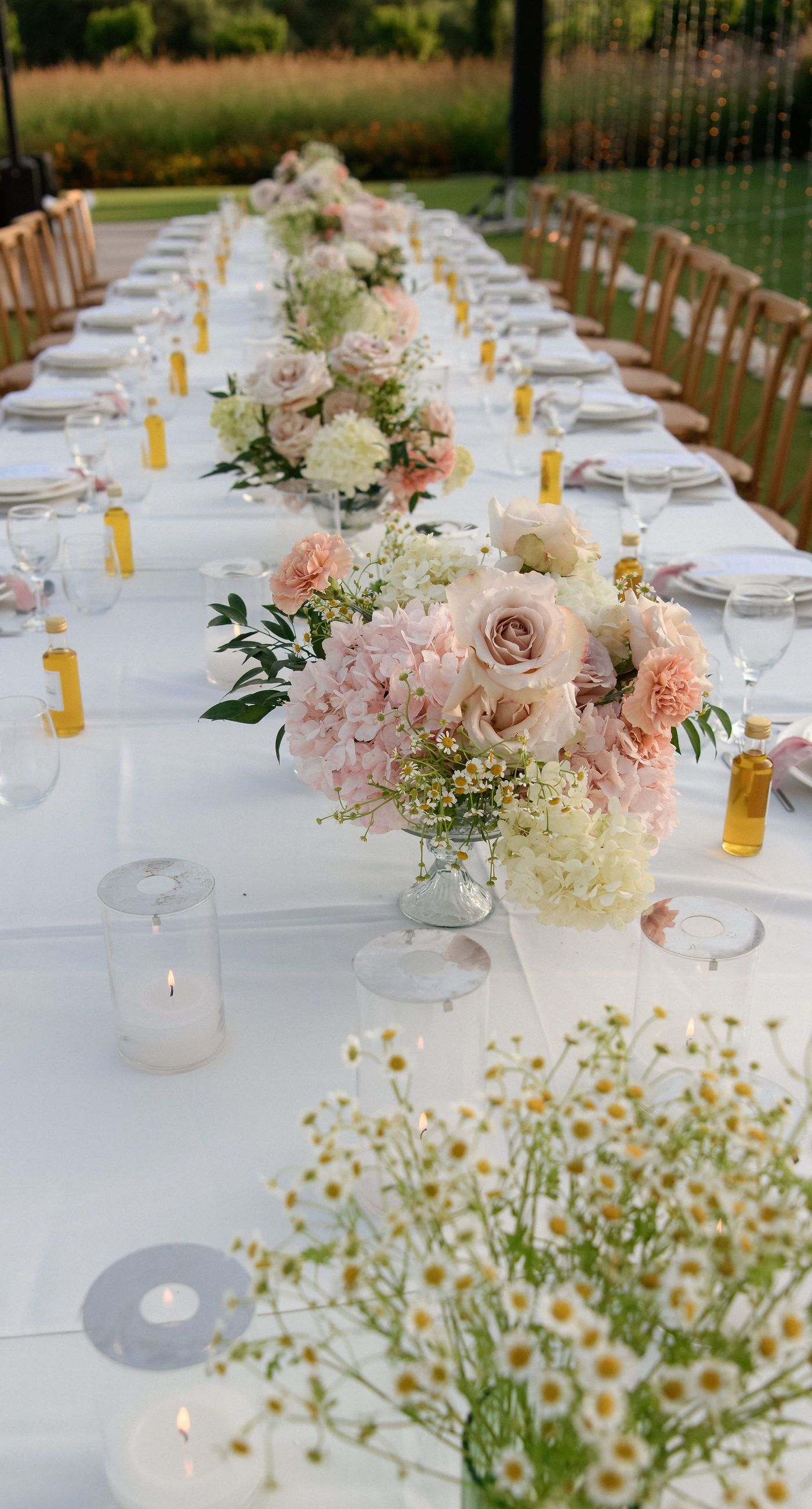 Sandra & Maximilian - In a serene garden setting, a couple stands embraced, surrounded by a breathtaking floral display. The bride's off-the-shoulder lace gown complements the groom's classic black tuxedo, both exuding elegance amid the lush, natural backdrop. Pastel blooms of blush and cream, expertly arranged, create an arch that seamlessly blends with the verdant landscape. Soft pinks and whites, interspersed with touches of greenery, add a delicate yet sophisticated touch. Rolling hills and distant mountains frame the scene, enhancing the timeless beauty and tranquility of this picturesque moment. - Photo 12