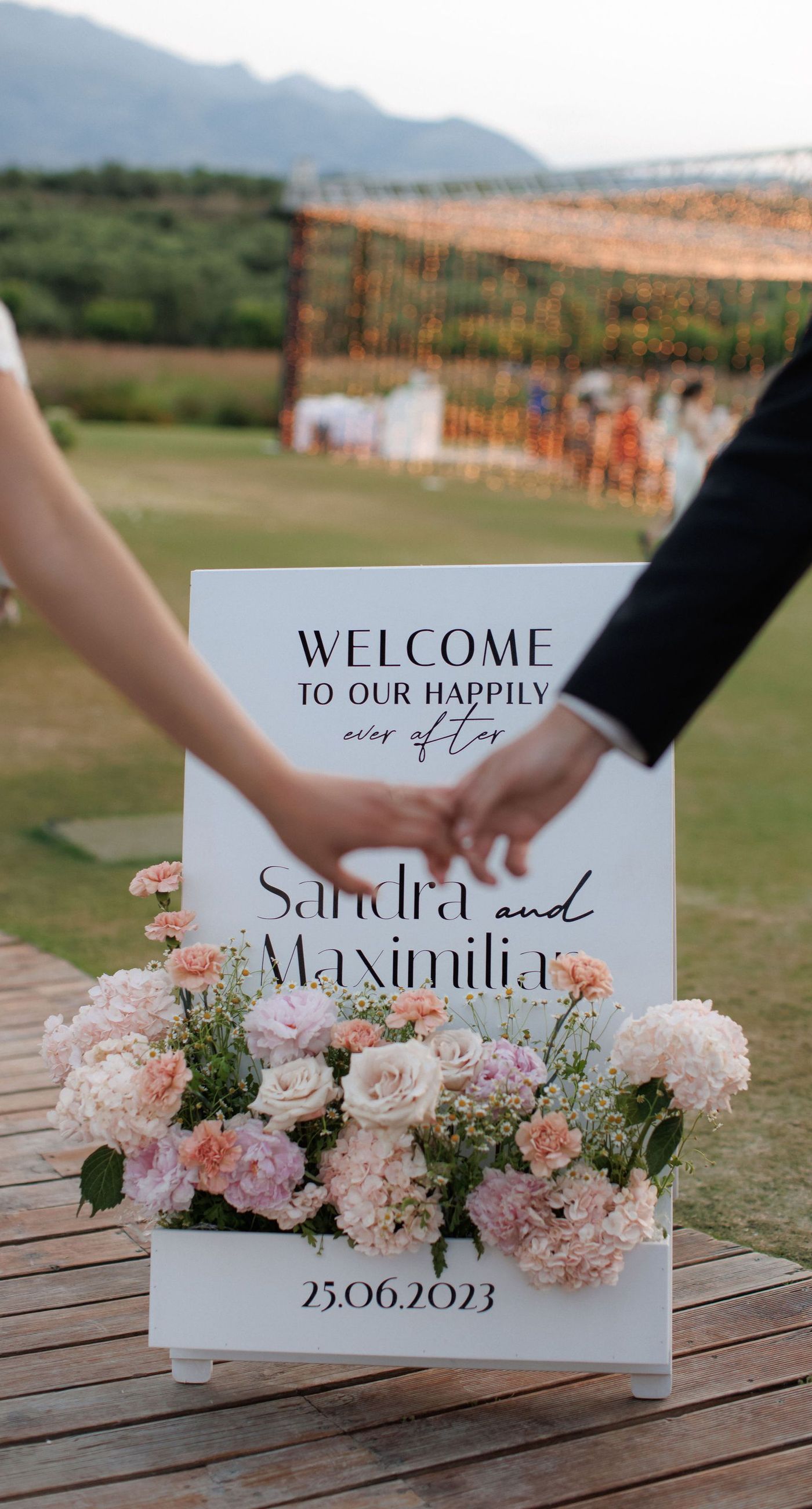 Sandra & Maximilian - In a serene garden setting, a couple stands embraced, surrounded by a breathtaking floral display. The bride's off-the-shoulder lace gown complements the groom's classic black tuxedo, both exuding elegance amid the lush, natural backdrop. Pastel blooms of blush and cream, expertly arranged, create an arch that seamlessly blends with the verdant landscape. Soft pinks and whites, interspersed with touches of greenery, add a delicate yet sophisticated touch. Rolling hills and distant mountains frame the scene, enhancing the timeless beauty and tranquility of this picturesque moment. - Photo 7