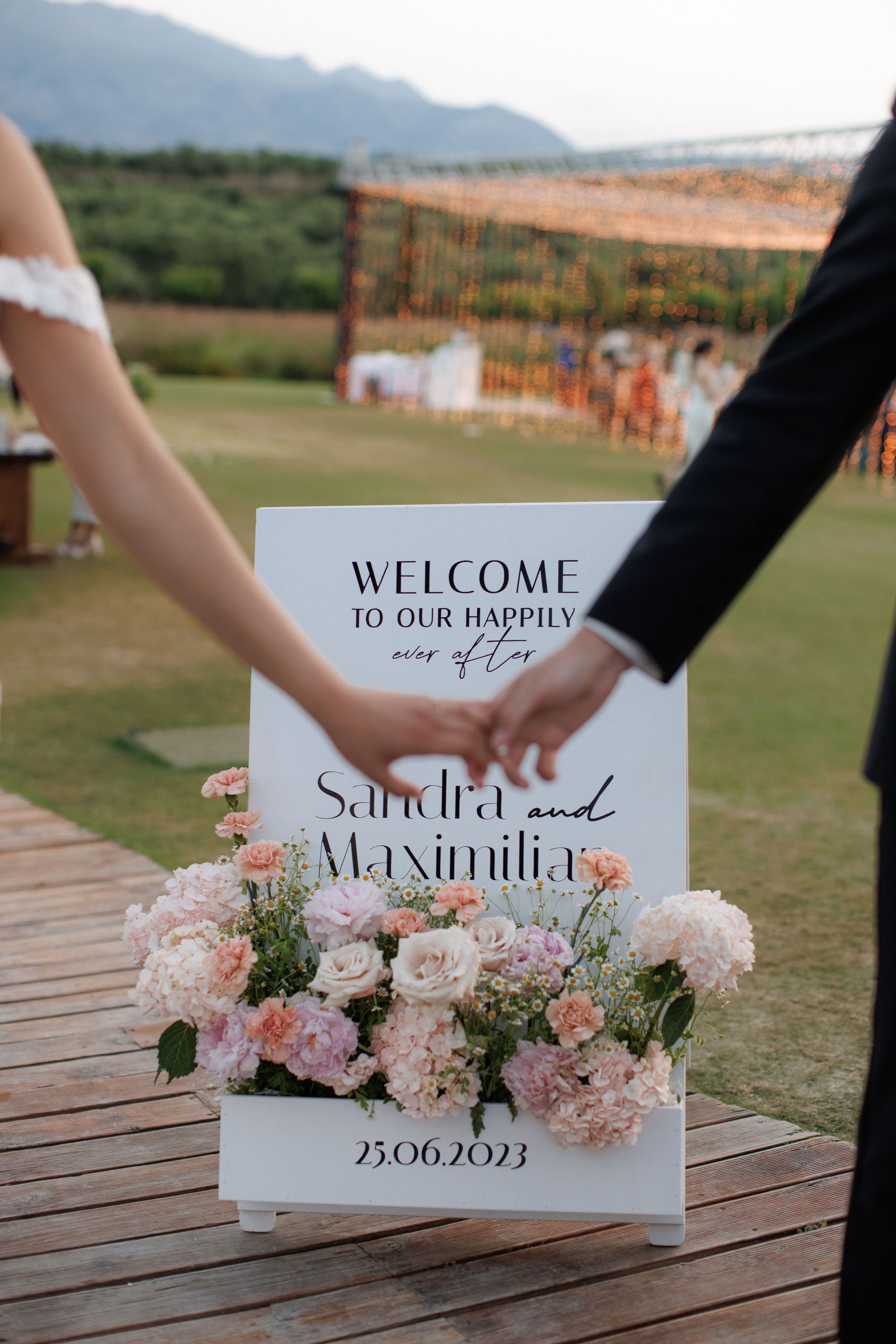 A bride and groom hold hands in front of a white welcome sign reading "Welcome to our happily ever after, Sandra and Maximilian," with a low arrangement of pale pink hydrangeas, peach carnations, blush roses, and white chamomile at its base. The bride's lace sleeve and the groom's dark suit sleeve frame the scene against a lawn softened by warm string lights, all part of wedding flowers at the Roxani estate in Greece.