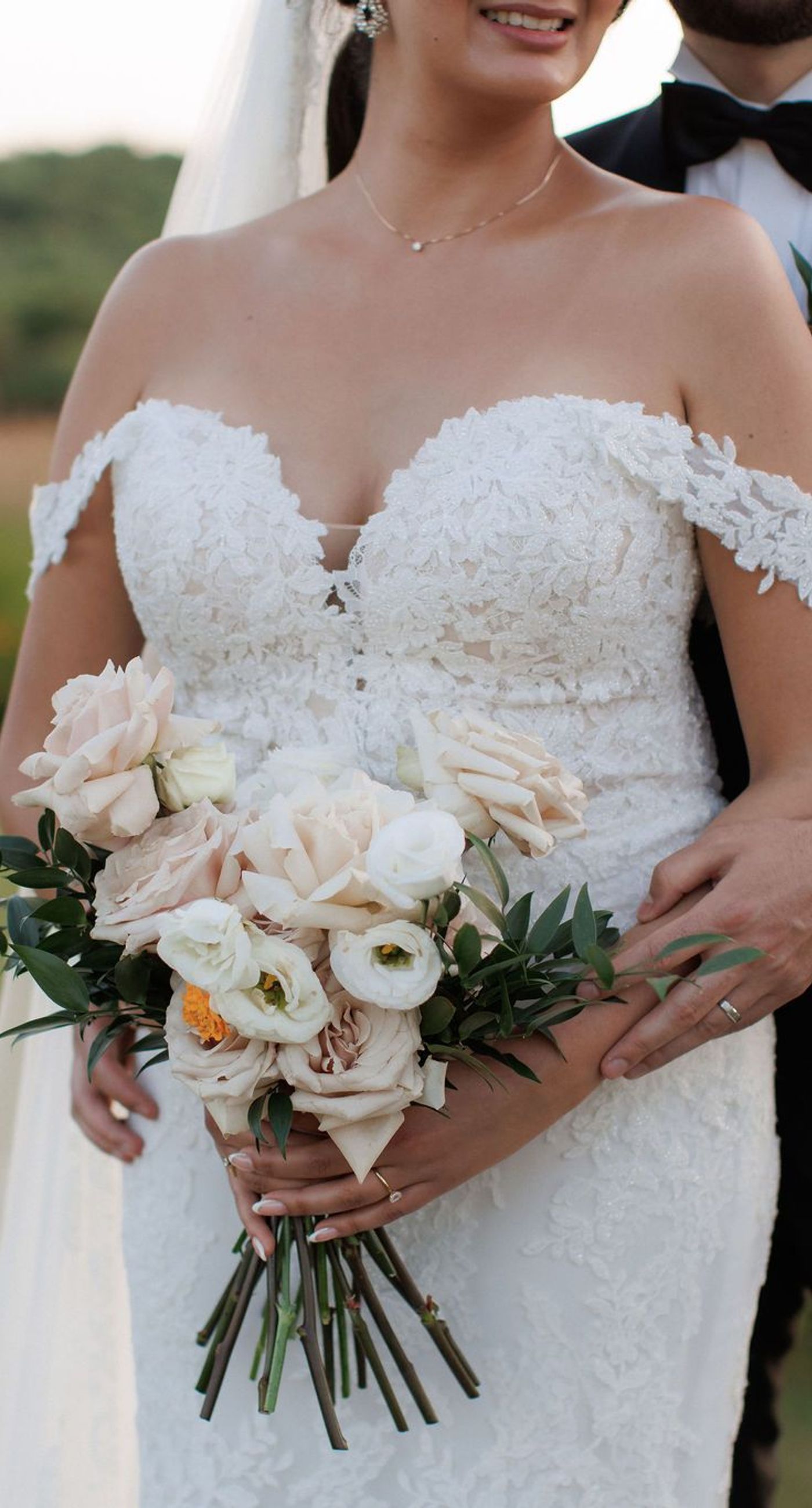 Sandra & Maximilian - In a serene garden setting, a couple stands embraced, surrounded by a breathtaking floral display. The bride's off-the-shoulder lace gown complements the groom's classic black tuxedo, both exuding elegance amid the lush, natural backdrop. Pastel blooms of blush and cream, expertly arranged, create an arch that seamlessly blends with the verdant landscape. Soft pinks and whites, interspersed with touches of greenery, add a delicate yet sophisticated touch. Rolling hills and distant mountains frame the scene, enhancing the timeless beauty and tranquility of this picturesque moment. - Photo 6
