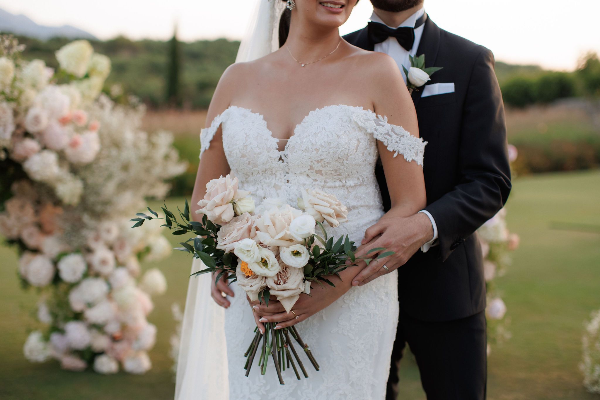 A bride in an off-the-shoulder lace gown holds a loose wedding bouquet of blush garden roses, white lisianthus, a small orange bloom and trailing green olive foliage, while a groom in a black tuxedo and bow tie stands behind her with his arm around her waist. Their wedding flowers are set against a soft, lush backdrop of white and blush blooms at Roxani estate in Greece, with green hills blurred in the evening light.