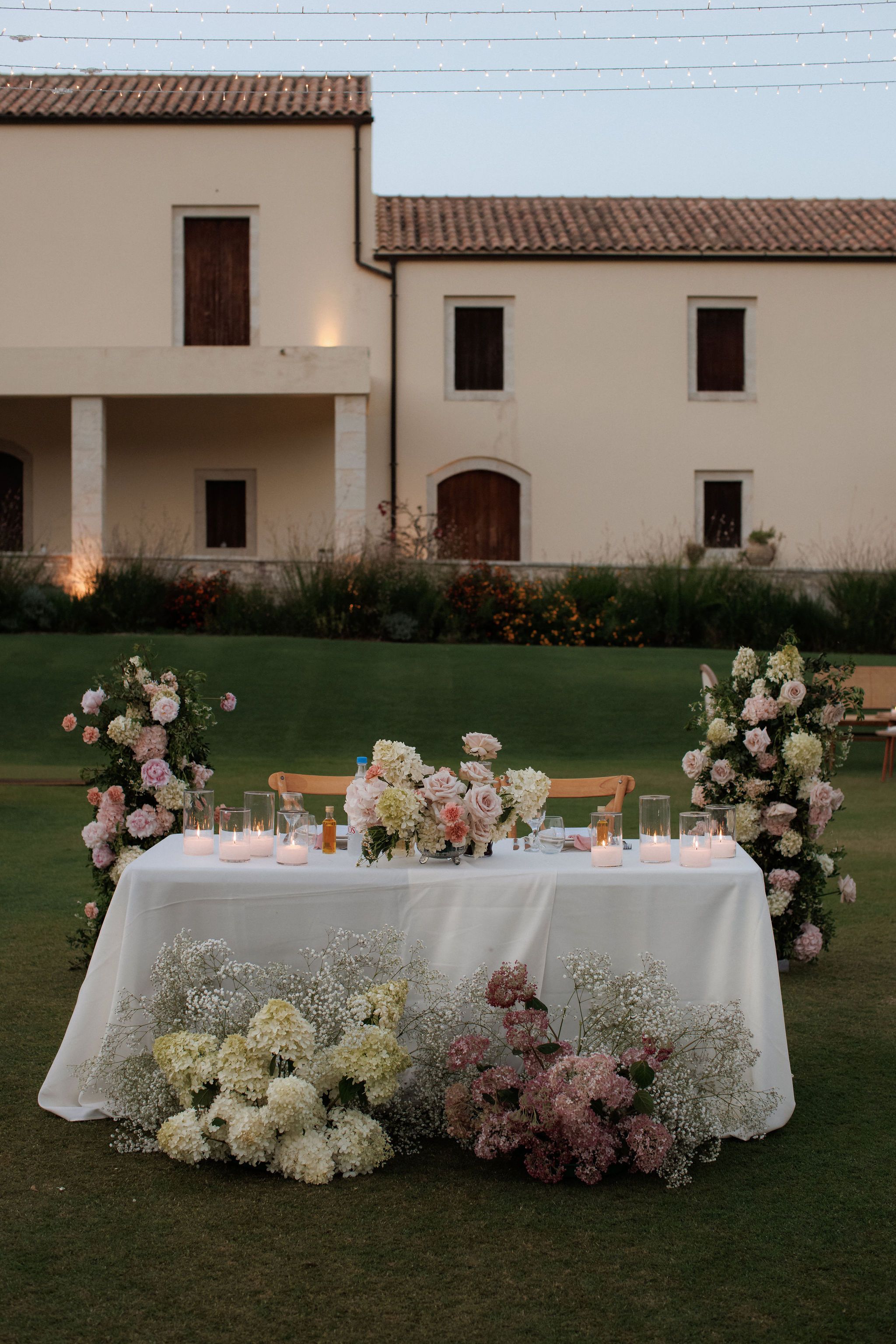 A long white-clothed sweetheart table sits on a lawn in front of a cream stone villa at dusk, dressed with tall pink and cream floral arches on either side and a low centrepiece of blush garden roses, peonies, cream hydrangeas and pale carnations. Glass hurricane candles glow along the table, and two wooden chairs face each other across it. This is a wedding table setting in Roxani estate in Greece, with soft pink and ivory tones and an evening mood.