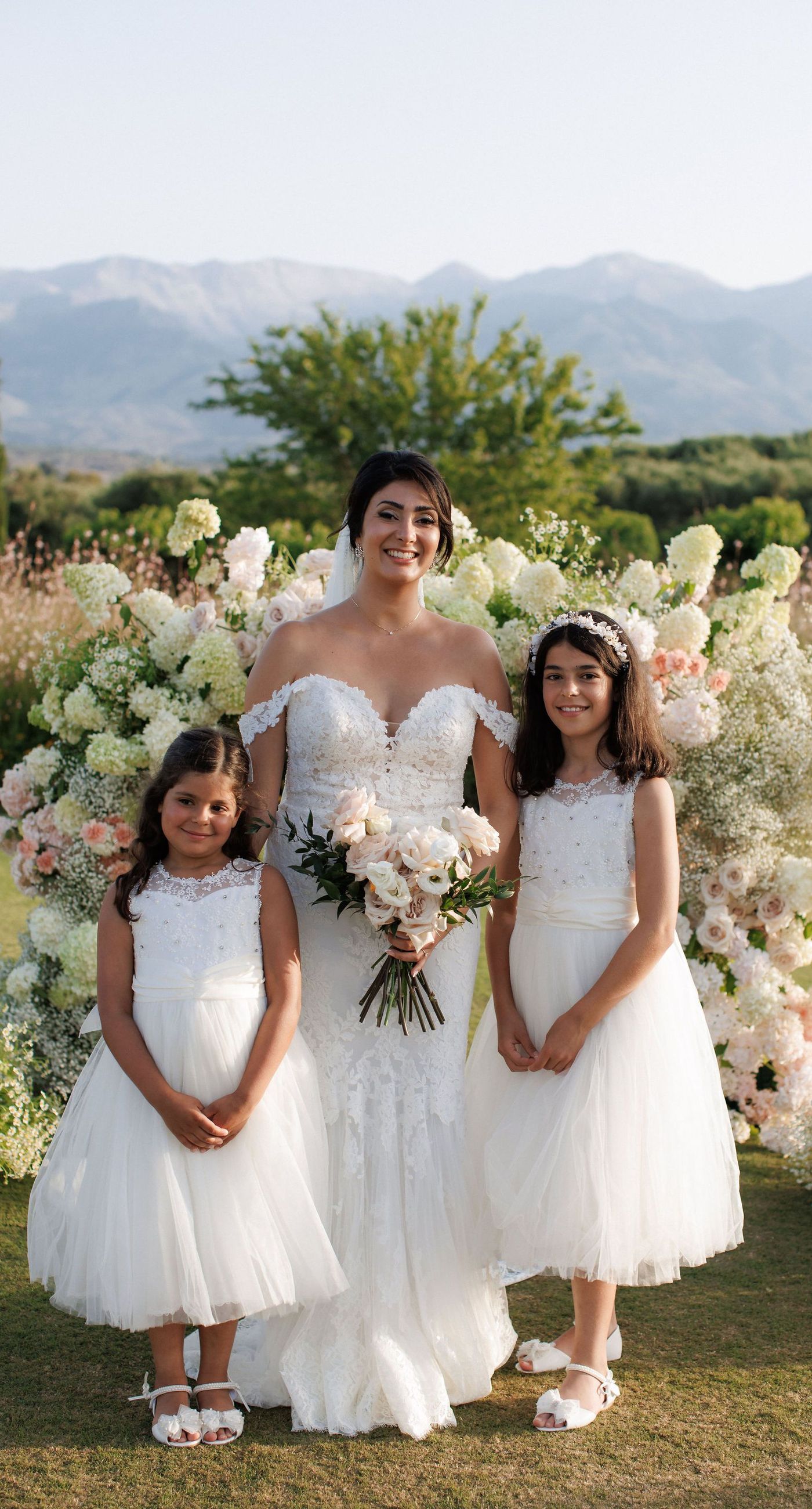 Sandra & Maximilian - In a serene garden setting, a couple stands embraced, surrounded by a breathtaking floral display. The bride's off-the-shoulder lace gown complements the groom's classic black tuxedo, both exuding elegance amid the lush, natural backdrop. Pastel blooms of blush and cream, expertly arranged, create an arch that seamlessly blends with the verdant landscape. Soft pinks and whites, interspersed with touches of greenery, add a delicate yet sophisticated touch. Rolling hills and distant mountains frame the scene, enhancing the timeless beauty and tranquility of this picturesque moment. - Photo 5