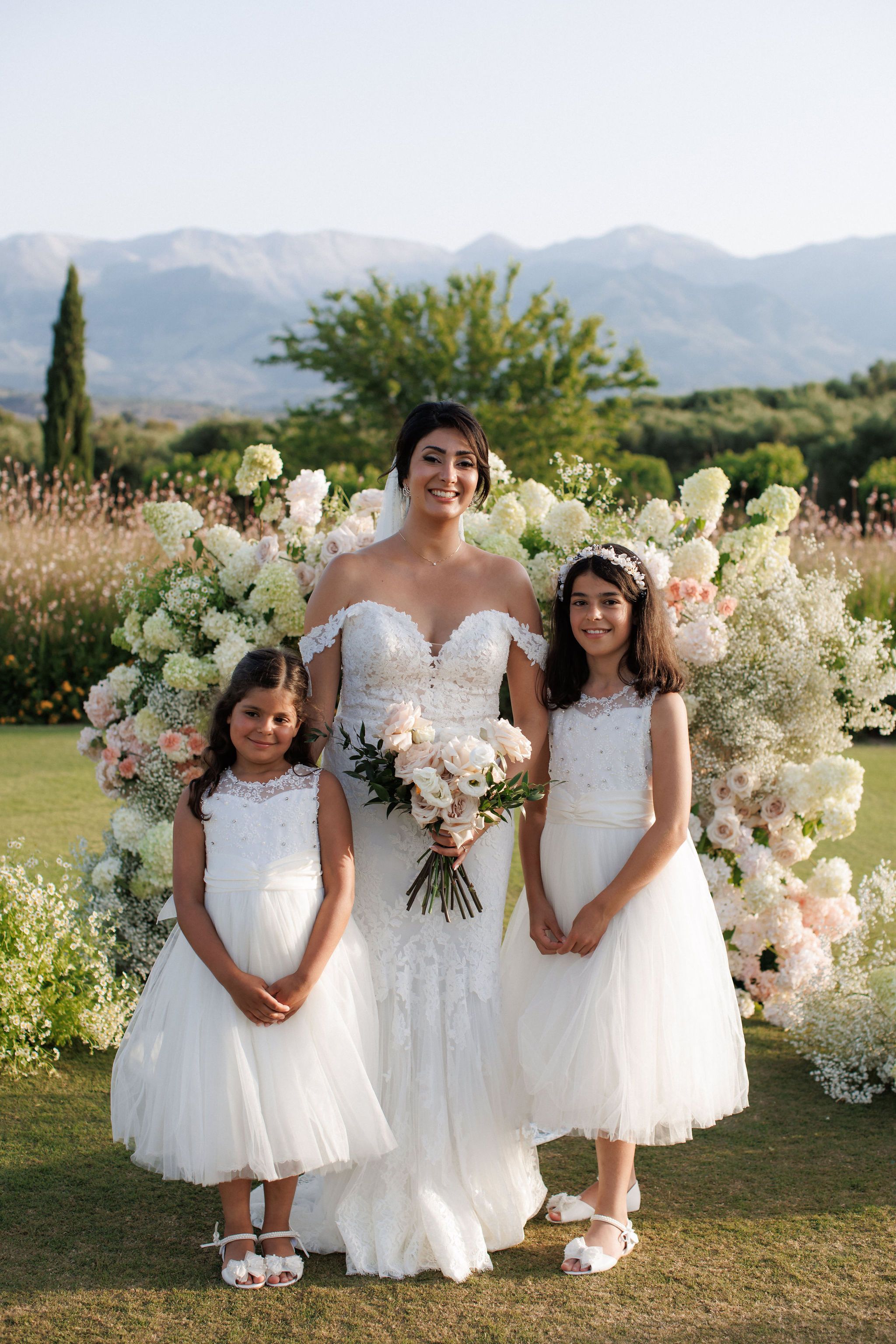 A bride in an off-the-shoulder lace gown stands smiling between two young flower girls in white tulle dresses, holding a soft bouquet of blush garden roses, ranunculus and white lisianthus with dark green foliage. Behind them is a lush arch of white hydrangeas, pale pink roses and baby's breath on a green lawn, with olive trees and mountains beyond. This is a wedding flower display at Roxani estate in Greece, captured in warm evening light.