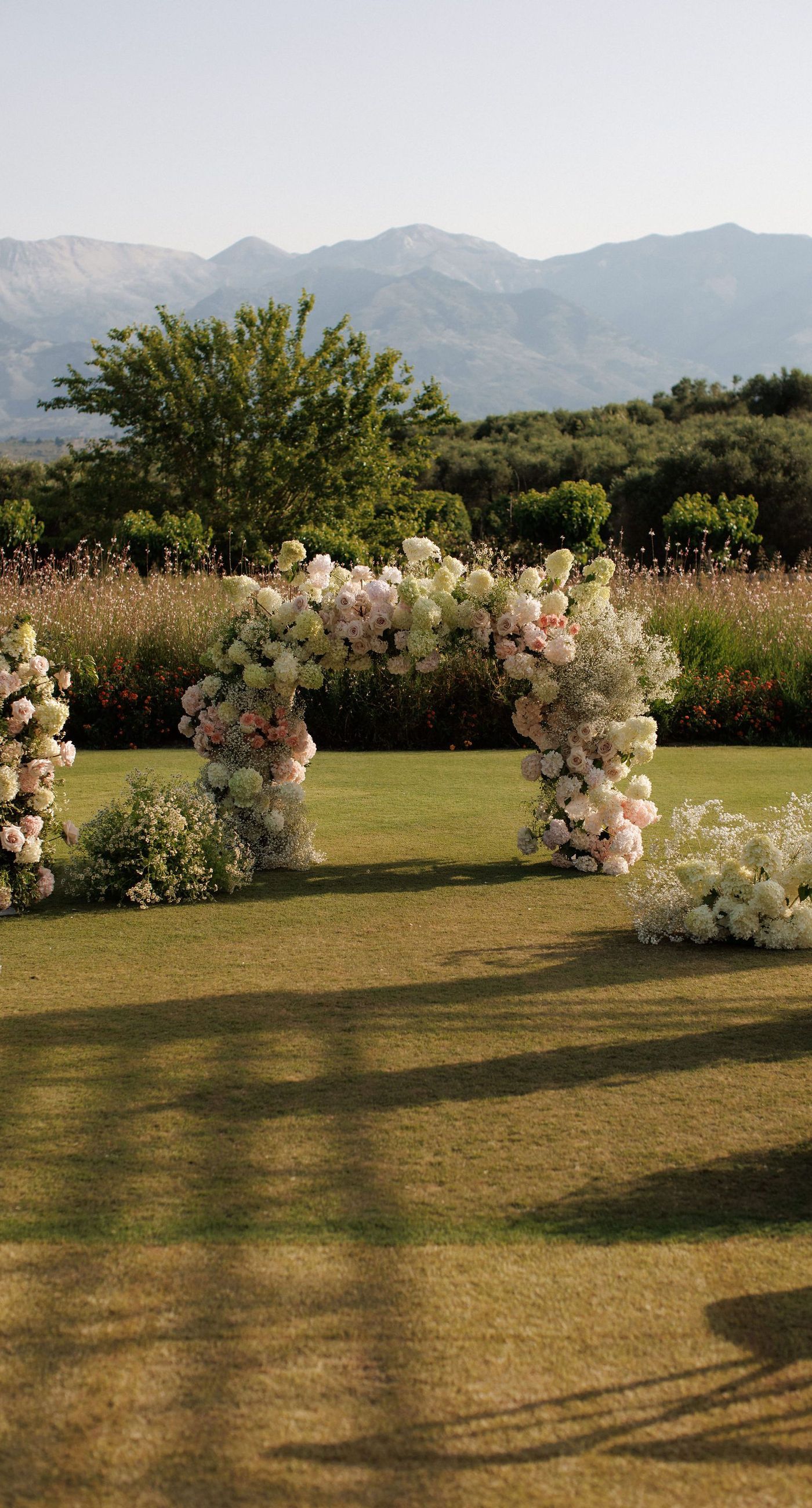 Sandra & Maximilian - In a serene garden setting, a couple stands embraced, surrounded by a breathtaking floral display. The bride's off-the-shoulder lace gown complements the groom's classic black tuxedo, both exuding elegance amid the lush, natural backdrop. Pastel blooms of blush and cream, expertly arranged, create an arch that seamlessly blends with the verdant landscape. Soft pinks and whites, interspersed with touches of greenery, add a delicate yet sophisticated touch. Rolling hills and distant mountains frame the scene, enhancing the timeless beauty and tranquility of this picturesque moment. - Photo 1