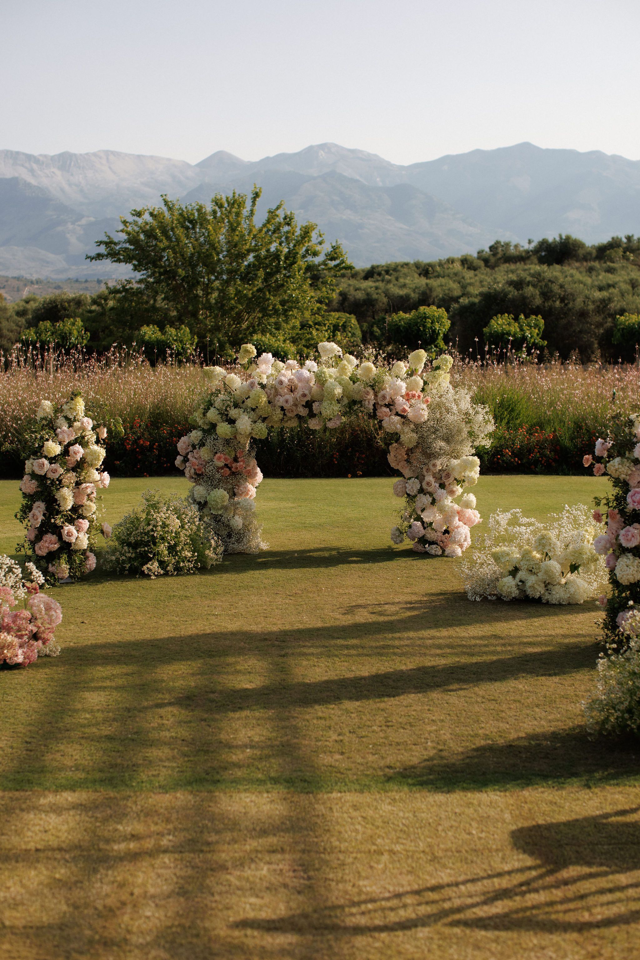 A wedding ceremony arch on a lawn at Roxani estate in Greece, lavishly covered in cream and blush garden roses, white and pale green hydrangeas, and sprays of baby's breath. Smaller floral columns flank the arch, and low mounds of white blooms sit on the grass, all set against a backdrop of tall pink-tinged grasses, olive trees and distant hills in soft golden evening light.