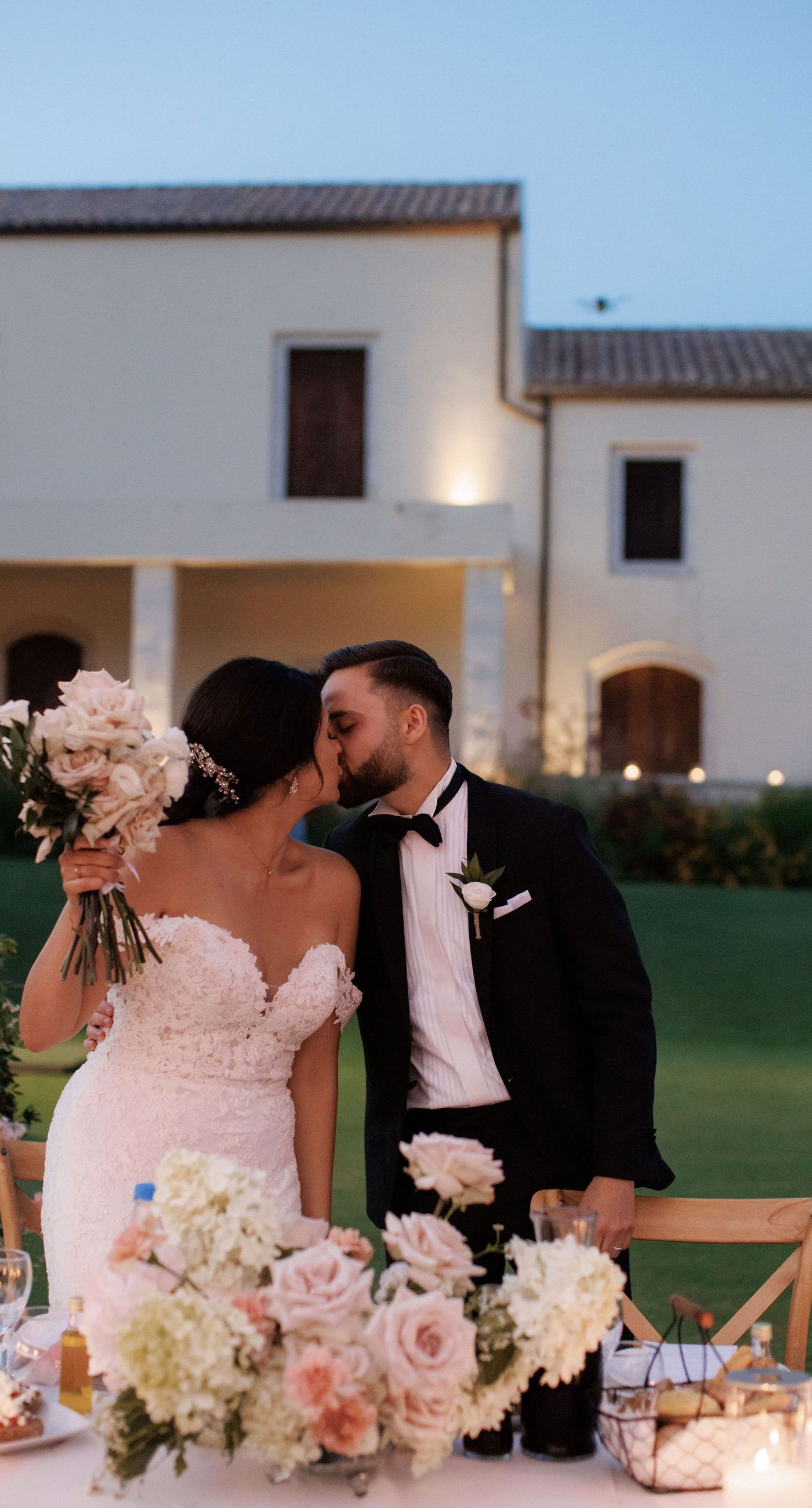 Sandra & Maximilian - In a serene garden setting, a couple stands embraced, surrounded by a breathtaking floral display. The bride's off-the-shoulder lace gown complements the groom's classic black tuxedo, both exuding elegance amid the lush, natural backdrop. Pastel blooms of blush and cream, expertly arranged, create an arch that seamlessly blends with the verdant landscape. Soft pinks and whites, interspersed with touches of greenery, add a delicate yet sophisticated touch. Rolling hills and distant mountains frame the scene, enhancing the timeless beauty and tranquility of this picturesque moment. - Photo 8