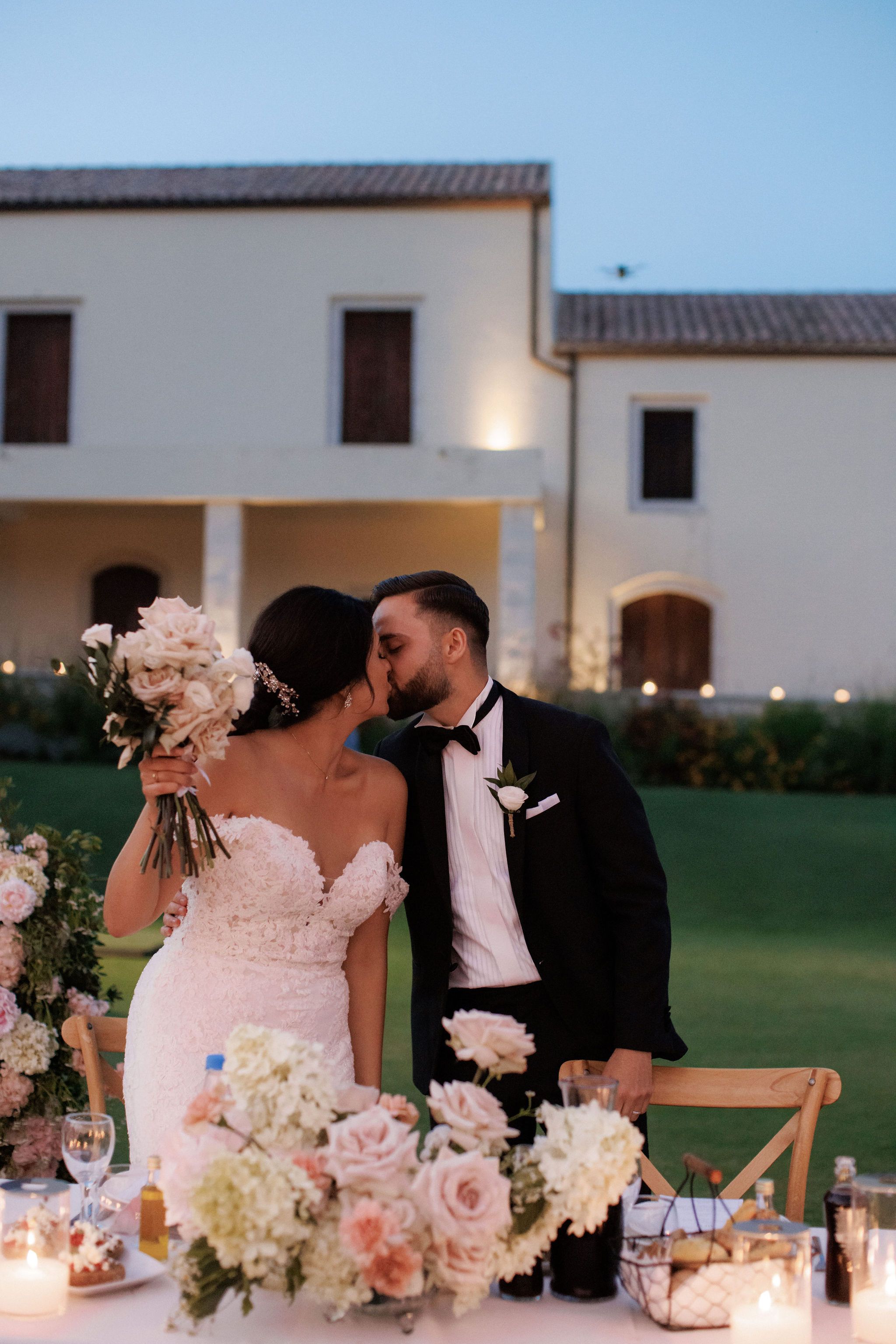 A bride in a fitted lace gown with a sweetheart neckline and a jewelled hair piece kisses her groom, who wears a black tuxedo, bow tie and a small white rose boutonniere, as she holds a pale blush bouquet of garden roses and foliage. In the foreground, a head table at the Roxani estate in Greece is dressed with blush roses, cream hydrangeas and candlelit glass hurricanes, with a lawn and a warmly lit stone building behind them at dusk. This is a wedding in Greece by manfler.