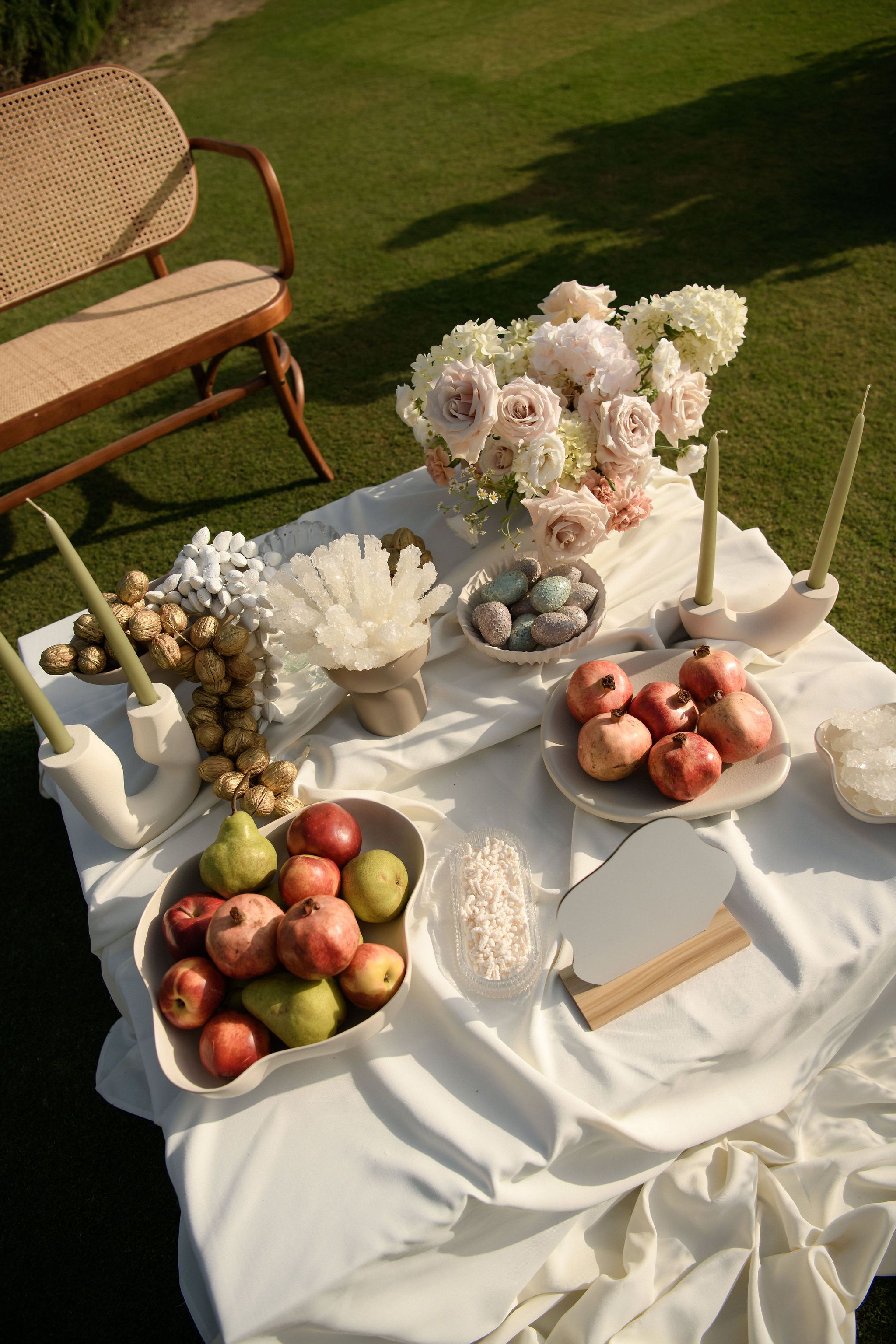A sunlit table draped in white satin on a lawn, styled for a wedding at Roxani estate in Greece. Pale pink garden roses and white blooms rise in a loose arrangement at the top, flanked by sage-green taper candles in curved cream holders. Below sit bowls of red apples, green pears and pomegranates, a dish of speckled blue-grey eggs, and a cluster of white crystal and gold-dusted walnuts, all in soft, earthy tones.