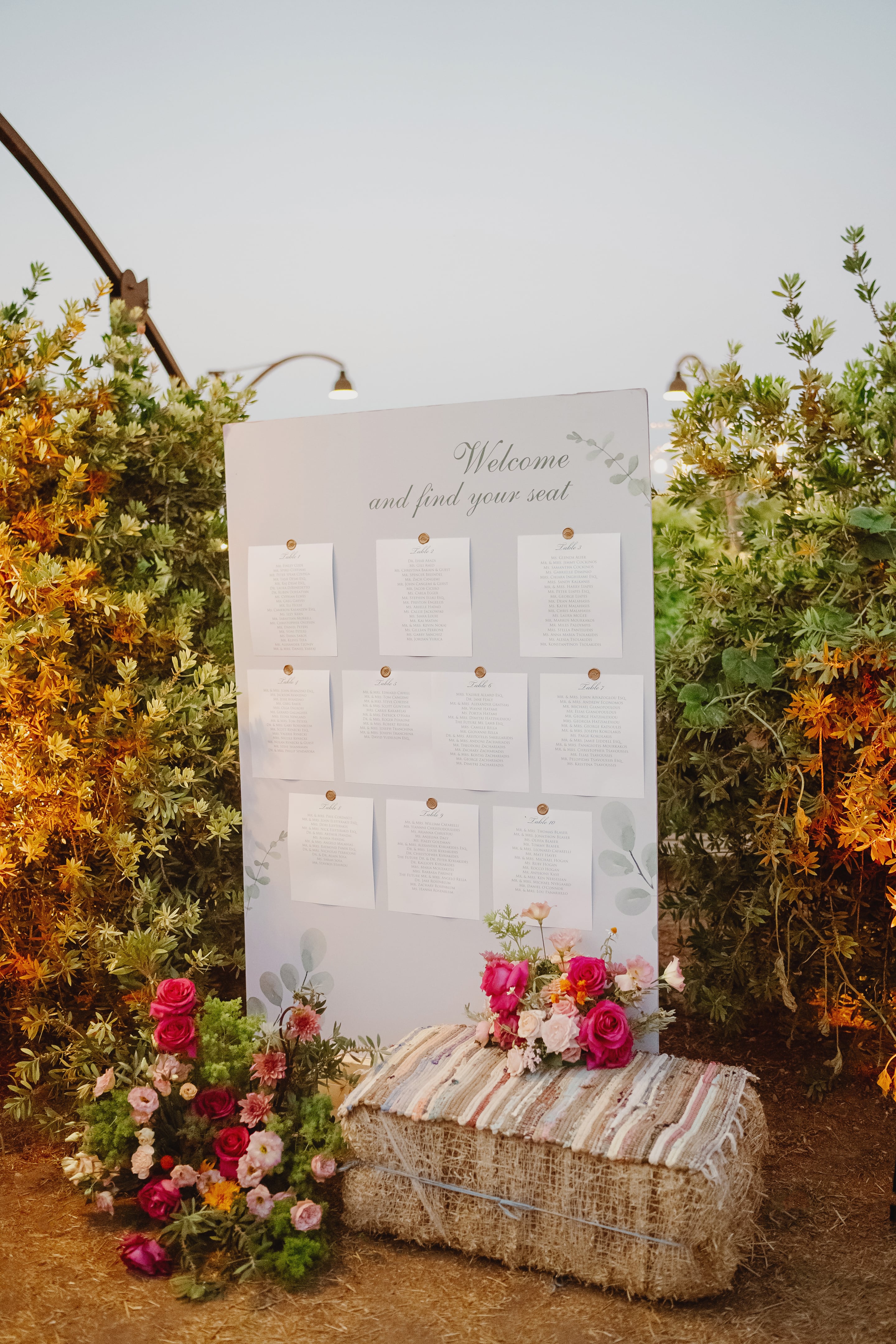 A wedding seating chart on a pale grey-blue board, with ten table lists pinned in a grid beneath the words "and find your seat" and decorated with sprigs of eucalyptus. In front sits a hay bale draped with a striped woven blanket, and on it and beside it are clusters of hot pink garden roses, blush lisianthus, pink dahlias and soft green foliage. Behind is a backdrop of golden-leaved shrubs in warm evening light, at a wedding in Greece.