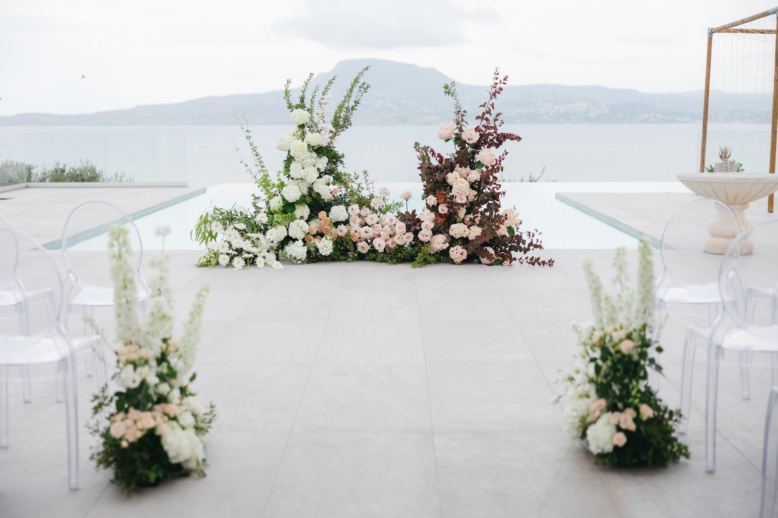 Wedding ceremony flowers at Villa Imperial in Greece: a low floral installation beside a pool, built from white hydrangeas, white daisies, blush garden roses and peach blooms, with tall dark plum foliage rising on the right and green stems on the left. Two matching arrangements of white delphinium and blush roses flank a pale stone aisle that leads toward the sea, and a hazy mountain island rises behind the calm water under a soft, overcast sky.