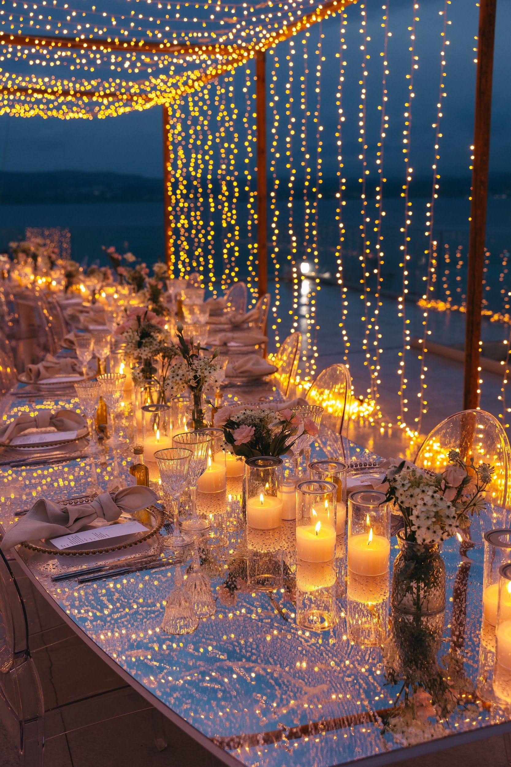 A long wedding dinner table at dusk at Villa Imperial in Greece, dressed with pillar candles in glass hurricanes, small vases of white and blush-pink blooms, and linen napkins tied in bows on gold-rimmed plates. Strings of warm fairy lights hang behind the table over a dark sea, and the glossy tabletop reflects the glow of the candles and lights.