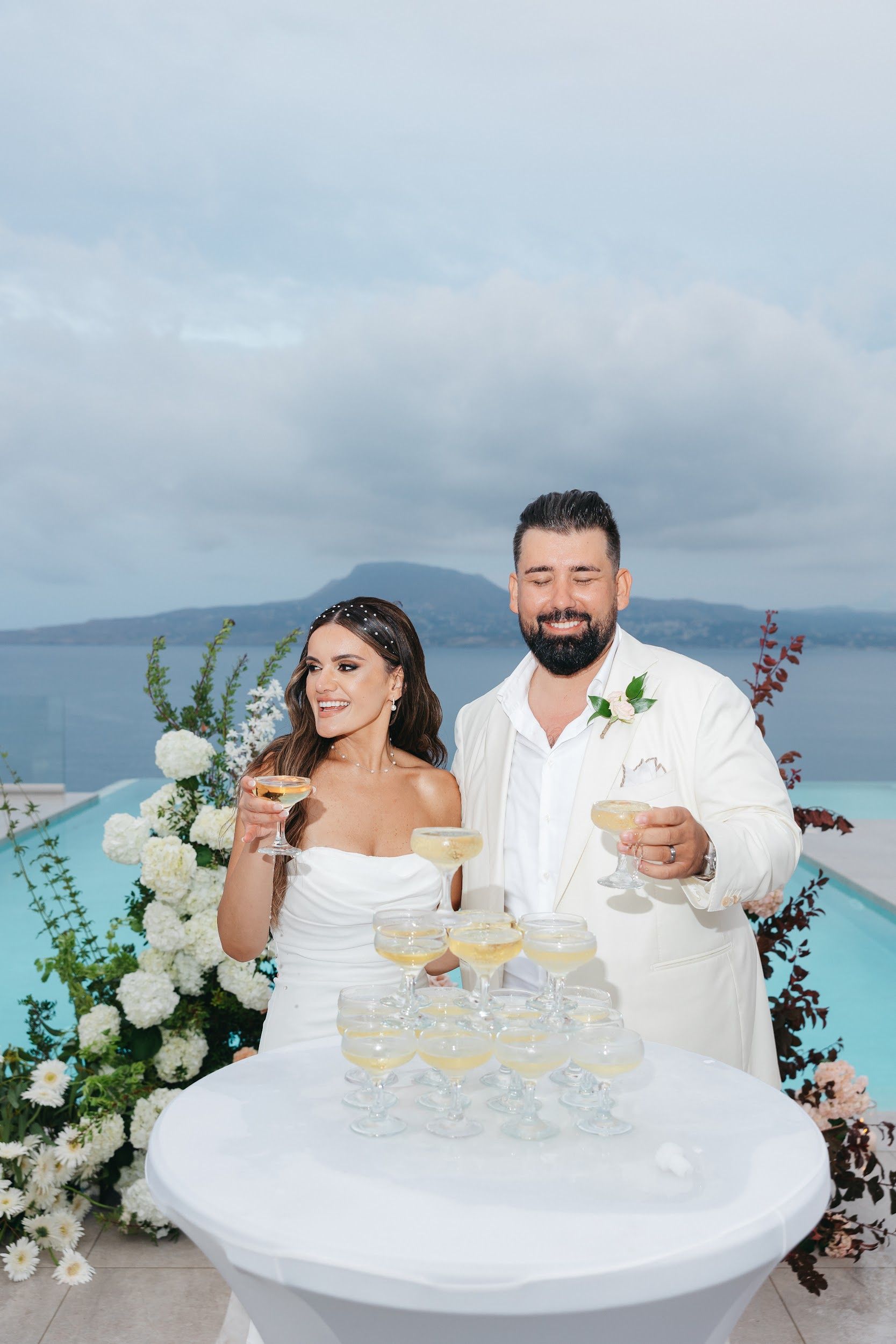 A bride and groom stand smiling beside a white-clothed cocktail table stacked with a pyramid of champagne coupes, each holding pale golden bubbly. Behind them, white hydrangeas, white daisies and trailing green foliage with dark burgundy stems frame an infinity pool that opens onto a calm sea and distant hills. The bride, in a strapless white gown with a pearl-trimmed headpiece, looks off to the side, glass raised, while the groom in a cream suit with a small white rose boutonniere raises his own glass. This is a wedding reception at Villa Imperial in Greece.