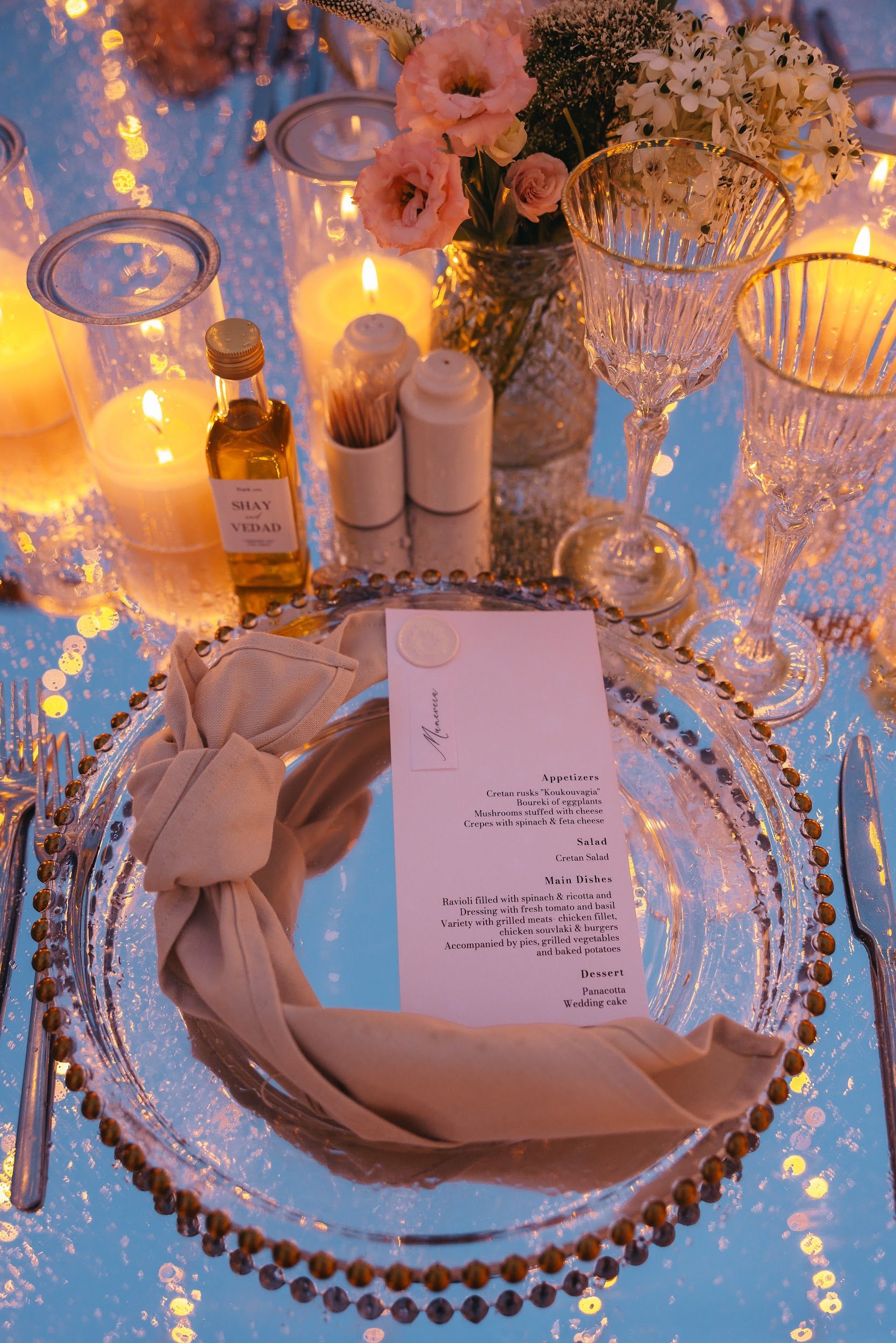 A close-up of a wedding place setting at Villa Imperial in Greece, lit by a row of warm pillar candles in glass hurricanes. A beaded glass charger holds a cream linen napkin knotted at the side and a printed menu card sealed with a wax disc, while a small bottle of olive oil labelled "Shay and Vedad" stands beside a toothpick holder and clear stemmed wine glasses with gold rims.