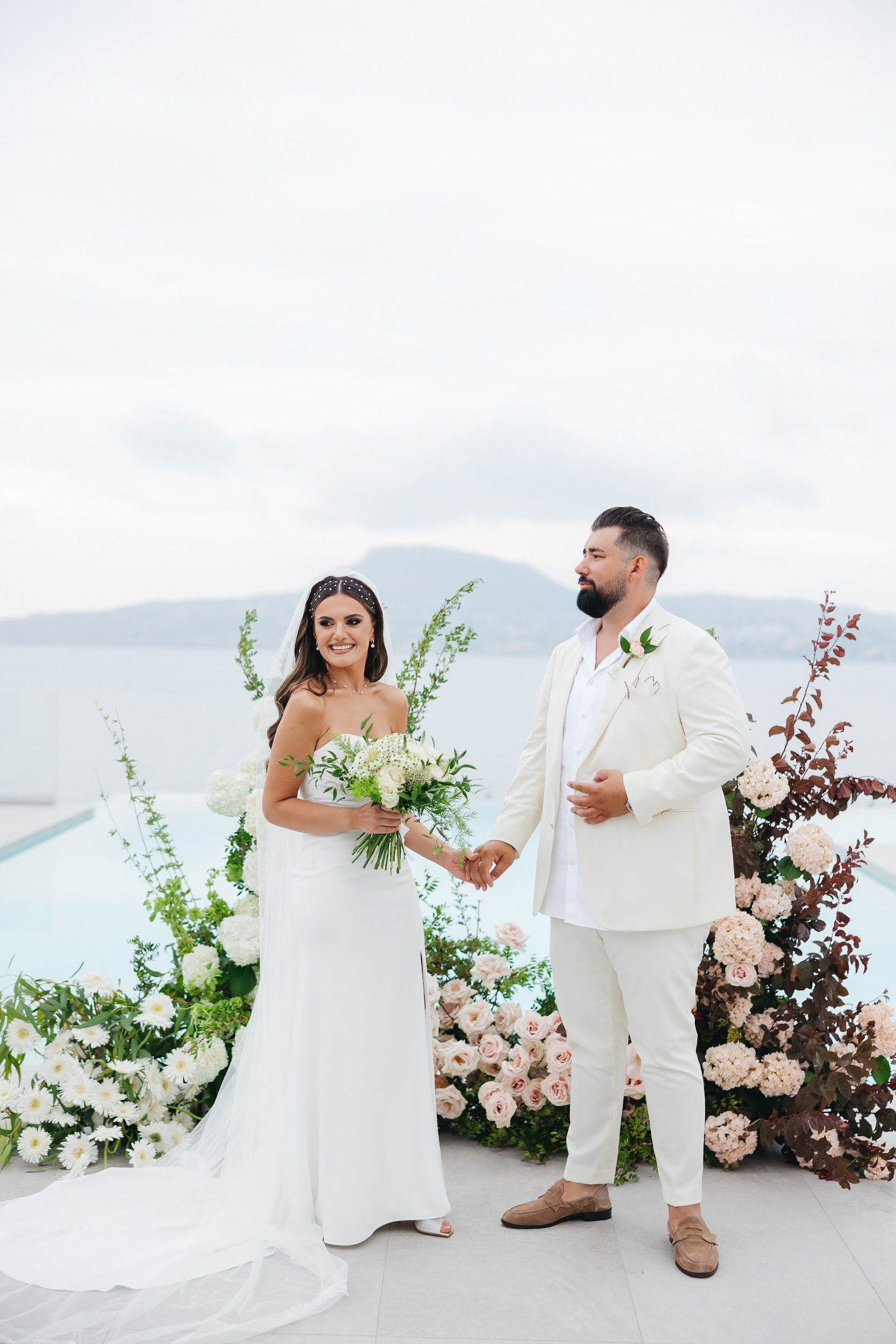 A bride in a strapless white gown with a long veil stands smiling beside her partner, who wears a cream suit and suede loafers, and they hold hands in front of a floral arch of white gerbera daisies, blush roses, pale hydrangeas and dark burgundy foliage beside an infinity pool at Villa Imperial in Greece. She carries a loose bouquet of white roses and green sprigs. The soft, pale palette and the sea behind them make the scene feel calm and airy.