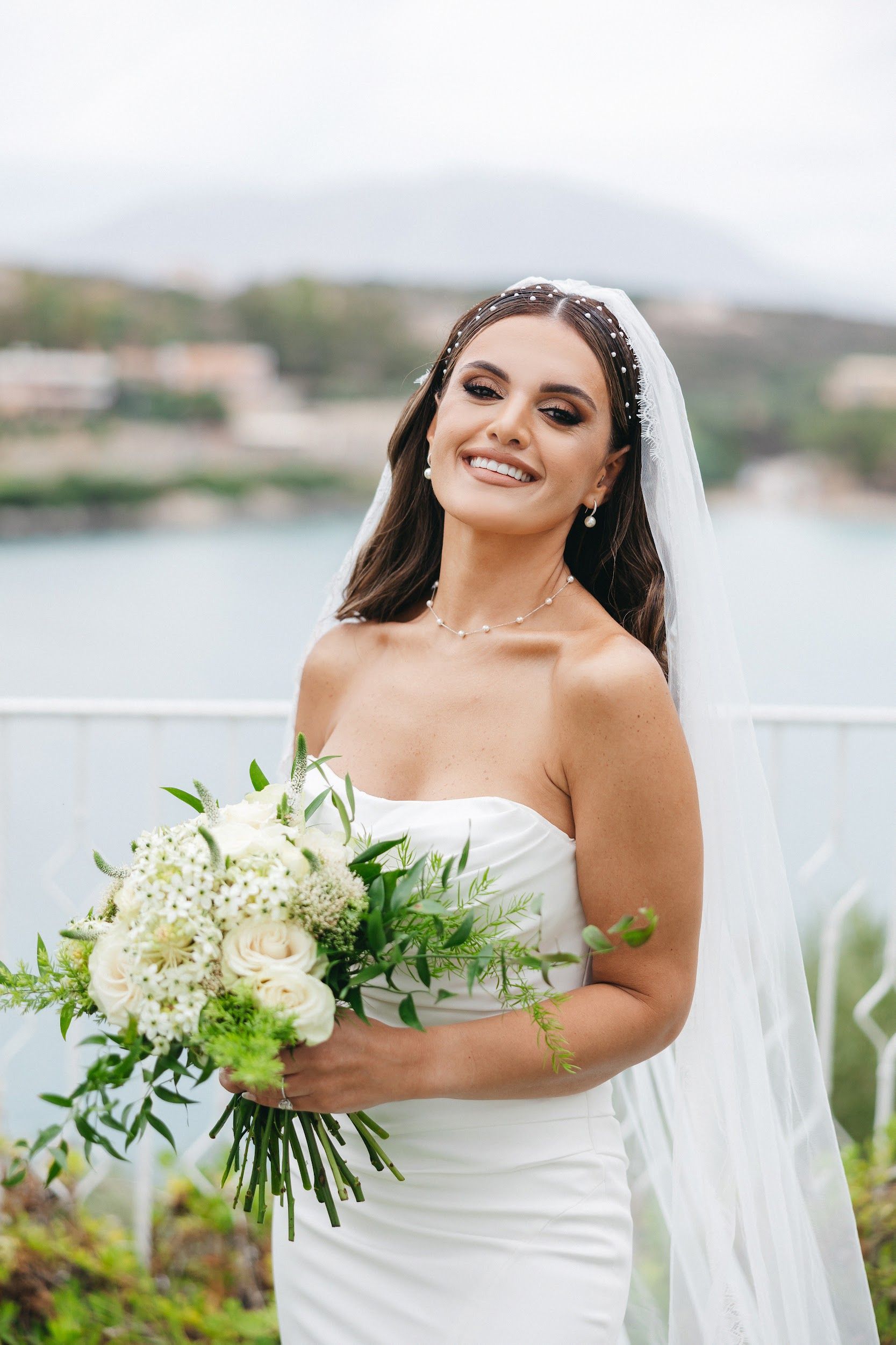 A smiling bride in a strapless white gown and a long, sheer veil stands on a terrace at Villa Imperial in Greece, holding a bridal bouquet of creamy white garden roses, white star-shaped blooms, feathery green foliage and sprigs of eucalyptus. Pearl-studded pins are tucked into her half-up hair, and she wears matching pearl earrings and a delicate pearl necklace. Behind her, a calm blue bay and soft green hills blur into a pale, airy background.