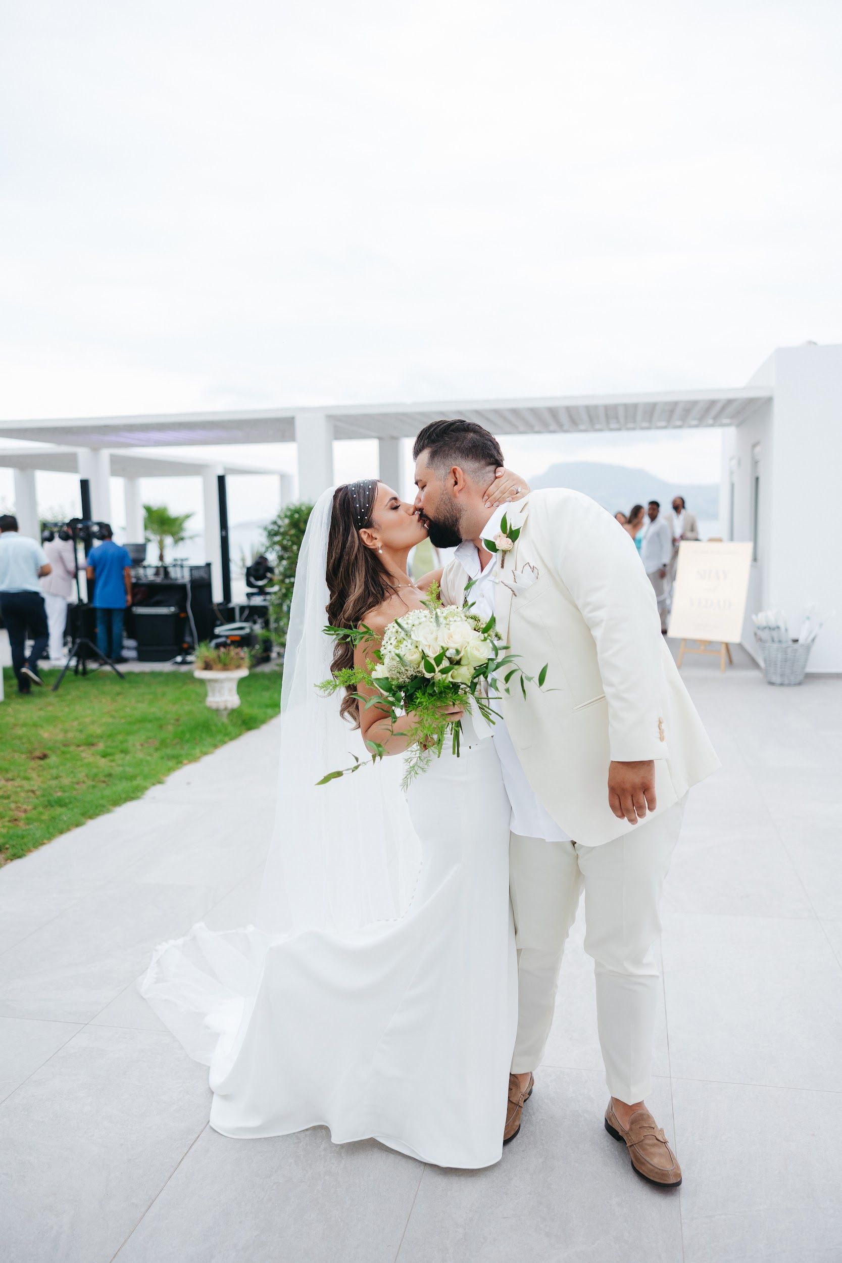 A bride and groom share a kiss on a white stone terrace at Villa Imperial in Greece, her bridal bouquet of white roses, hydrangea and trailing green foliage held in front of her. She wears a fitted white gown and a long veil with a pearl-trimmed headpiece, while he is dressed in a cream suit with a small white rose boutonniere. Behind them, white pergola beams and a pale sky frame the scene, with guests and a wooden welcome sign reading "Shay & Vedad" in the background.